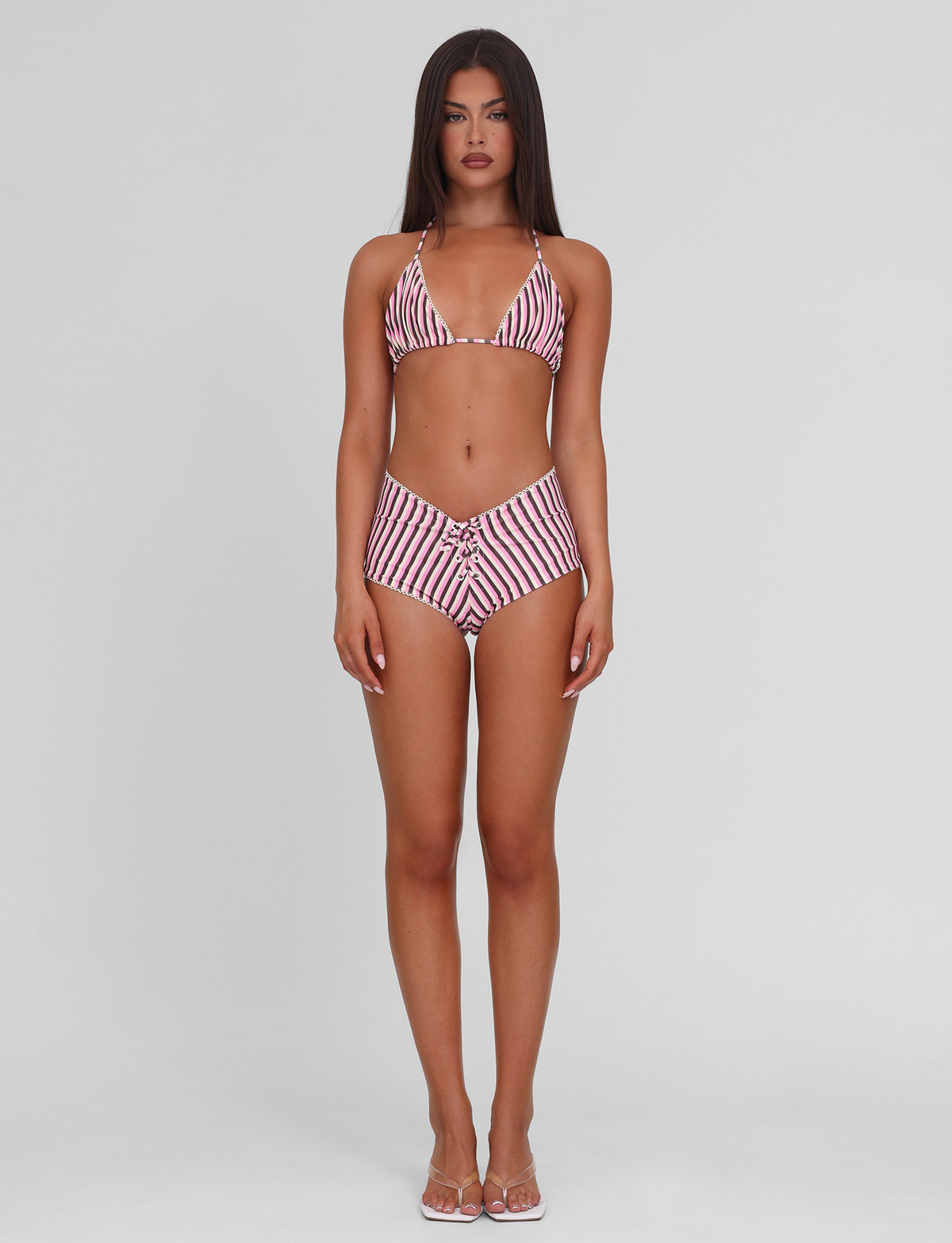 A woman stands facing forward, wearing the TIGERMIST DAZZLE BIKINI TOP - STRIPE with high-waisted bottoms and clear sandals, set against a plain light gray background.