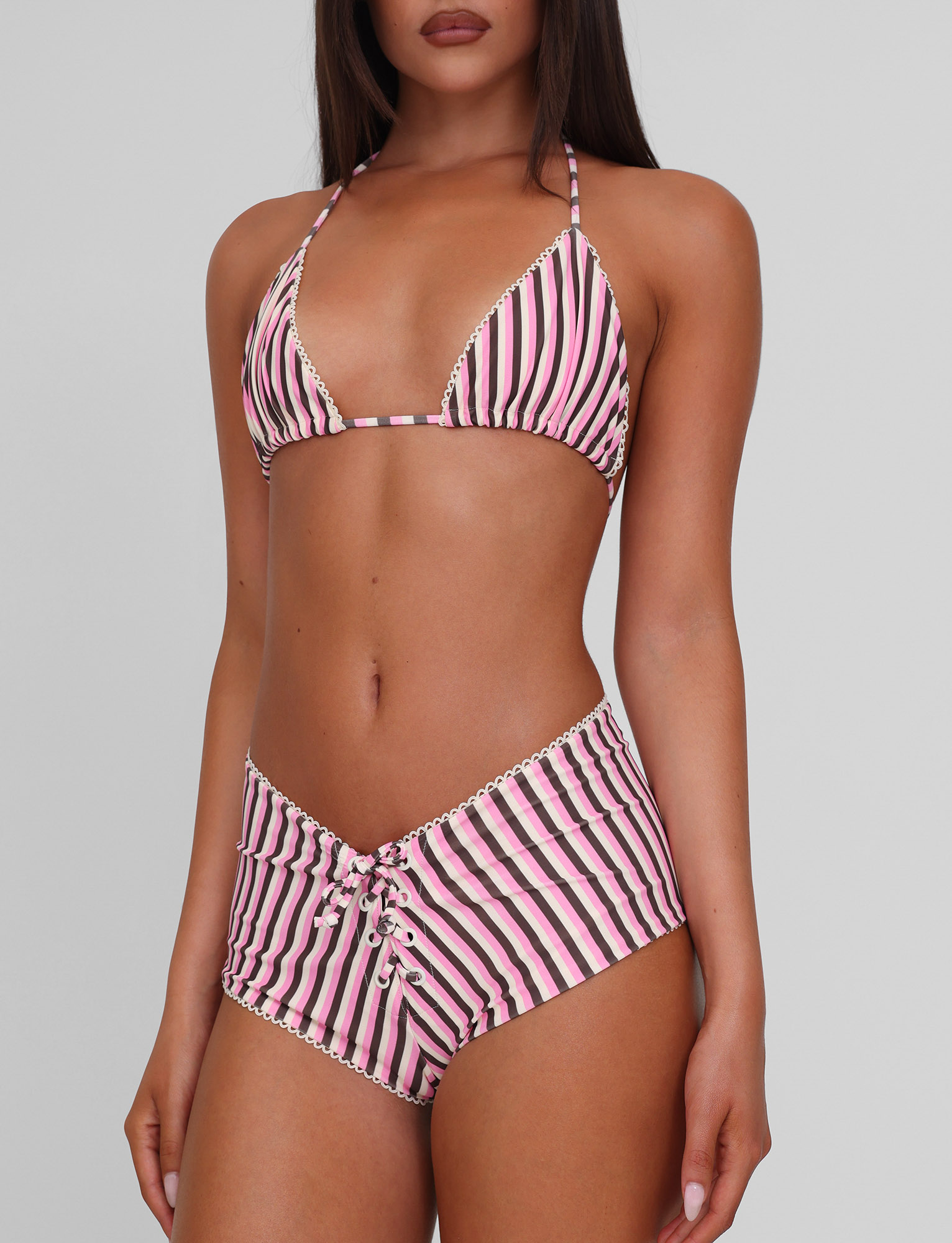 A woman models the TIGERMIST DAZZLE BIKINI TOP - STRIPE, a pink, white, and black high-stretch triangle bikini with lace-up, high-waisted bottoms, against a plain light gray background.