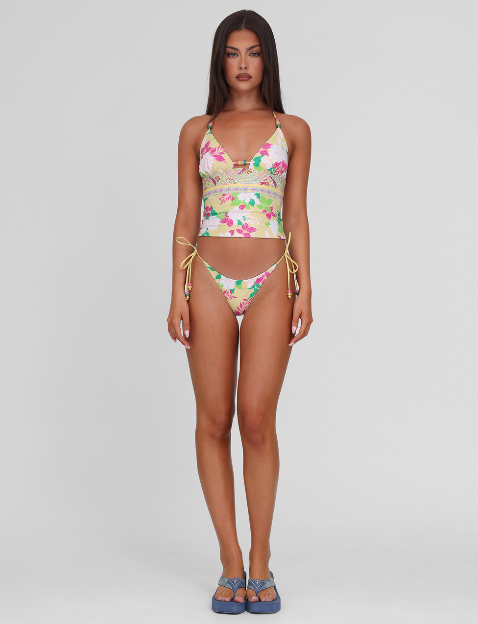 A woman with long brown hair stands against a plain background, wearing the TIGERMIST FINLEA TANKINI TOP in paisley border print and blue platform sandals. She faces forward with a neutral expression.