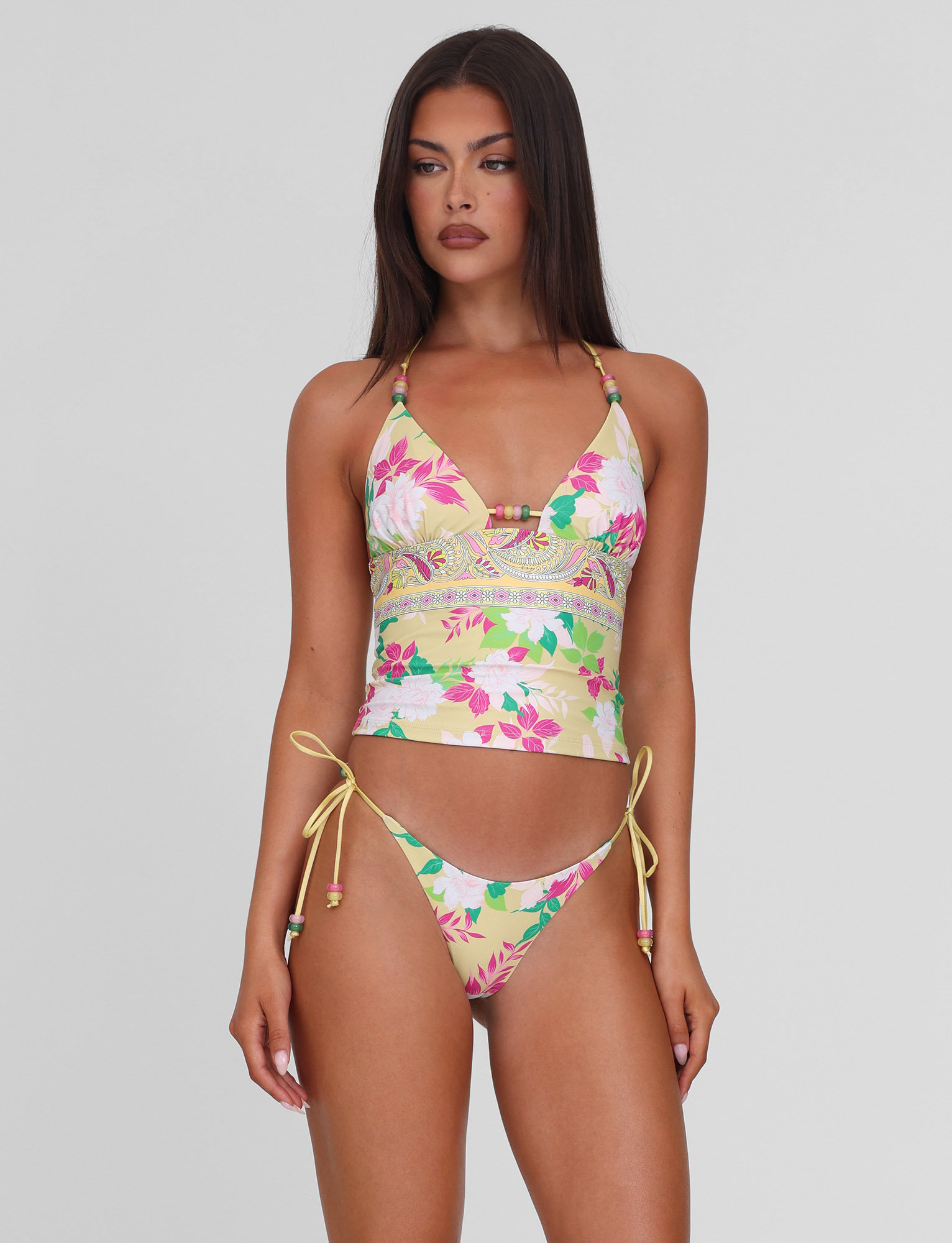 A woman with long brown hair wears the TIGERMIST FINLEA TANKINI TOP - PAISLEY BORDER and matching side-tie bikini bottoms in a yellow flower and paisley print, standing against a plain light gray background.