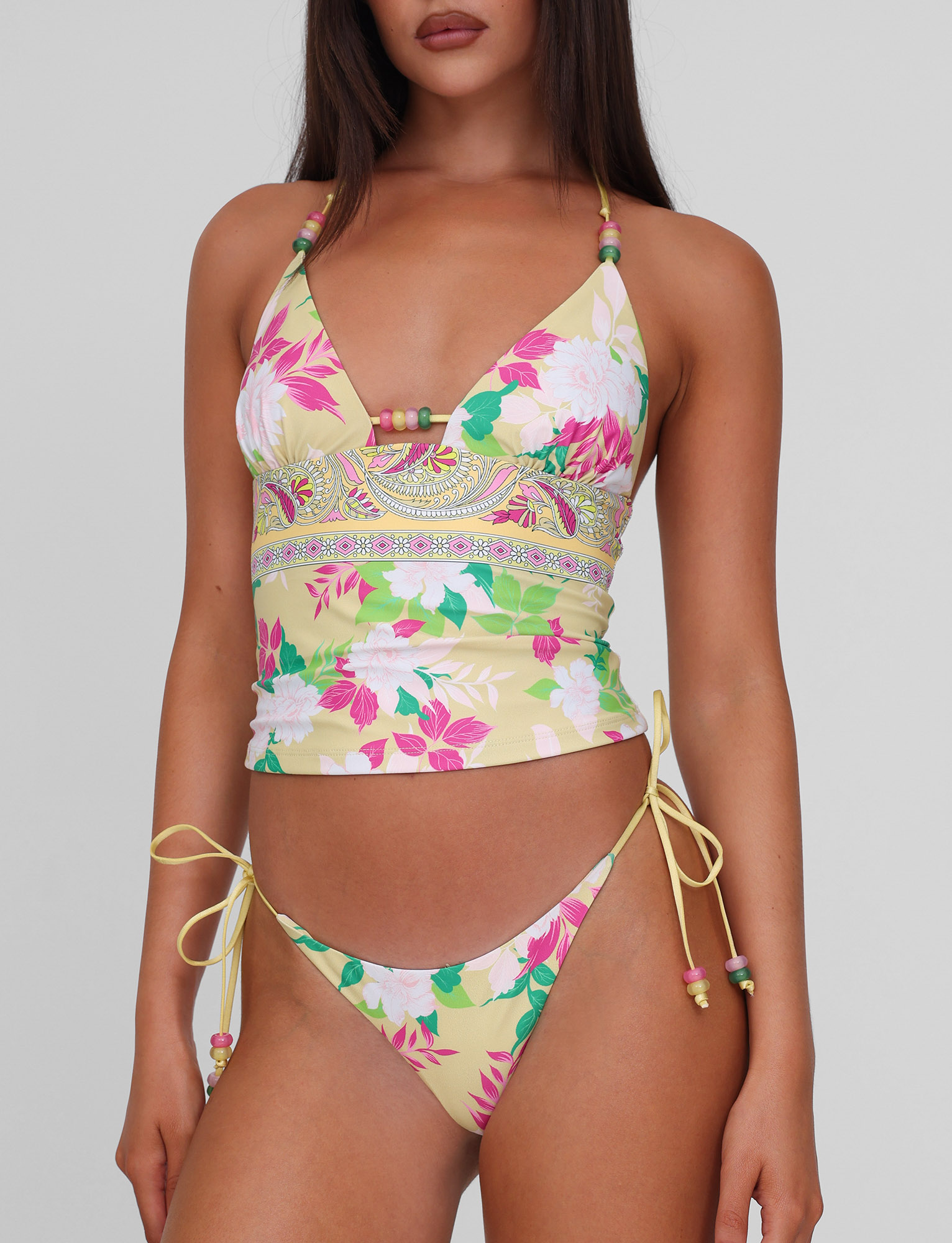 A woman wears the TIGERMIST FINLEA TANKINI TOP - PAISLEY BORDER, featuring yellow floral prints with pink, white, and green accents, paired with matching string bikini bottoms. The image is cropped from her shoulders to upper thighs.