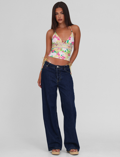A woman with straight dark hair wears the TIGERMIST FINLEA TANKINI TOP in paisley border print, featuring thin straps and drawstring sides, paired with high-waisted wide-leg dark blue jeans and platform sandals against a plain background.
