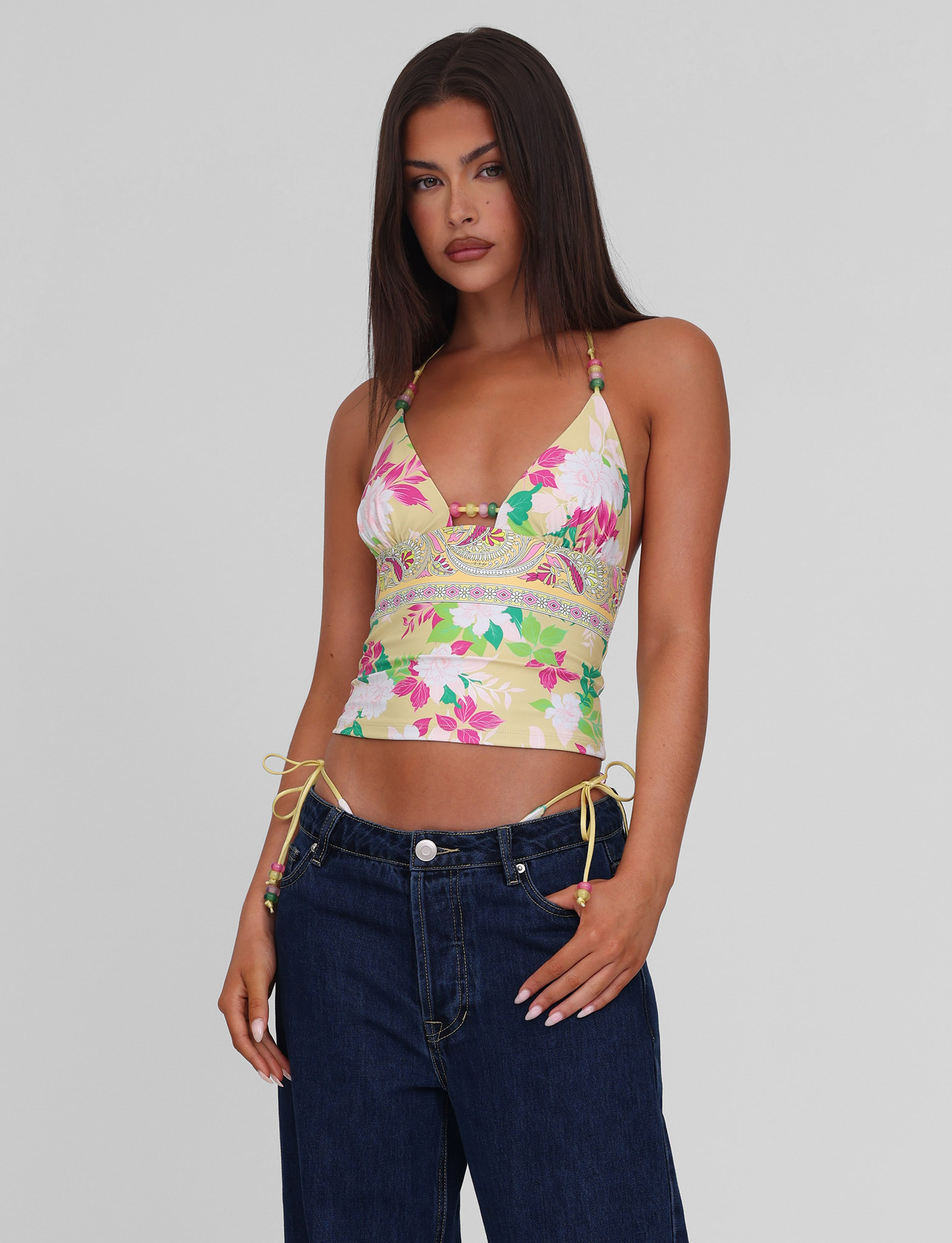 A woman with long dark hair wears the TIGERMIST FINLEA TANKINI TOP - PAISLEY BORDER with side ties and loose blue jeans, standing against a plain light gray background and facing the camera with a neutral expression.