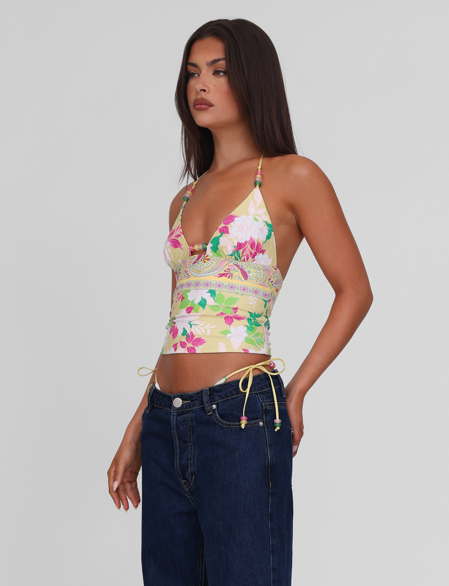 A woman with long dark hair models the TIGERMIST FINLEA TANKINI TOP - PAISLEY BORDER, paired with dark blue jeans, standing against a plain light gray background and looking slightly to the side with one hand in her pocket.