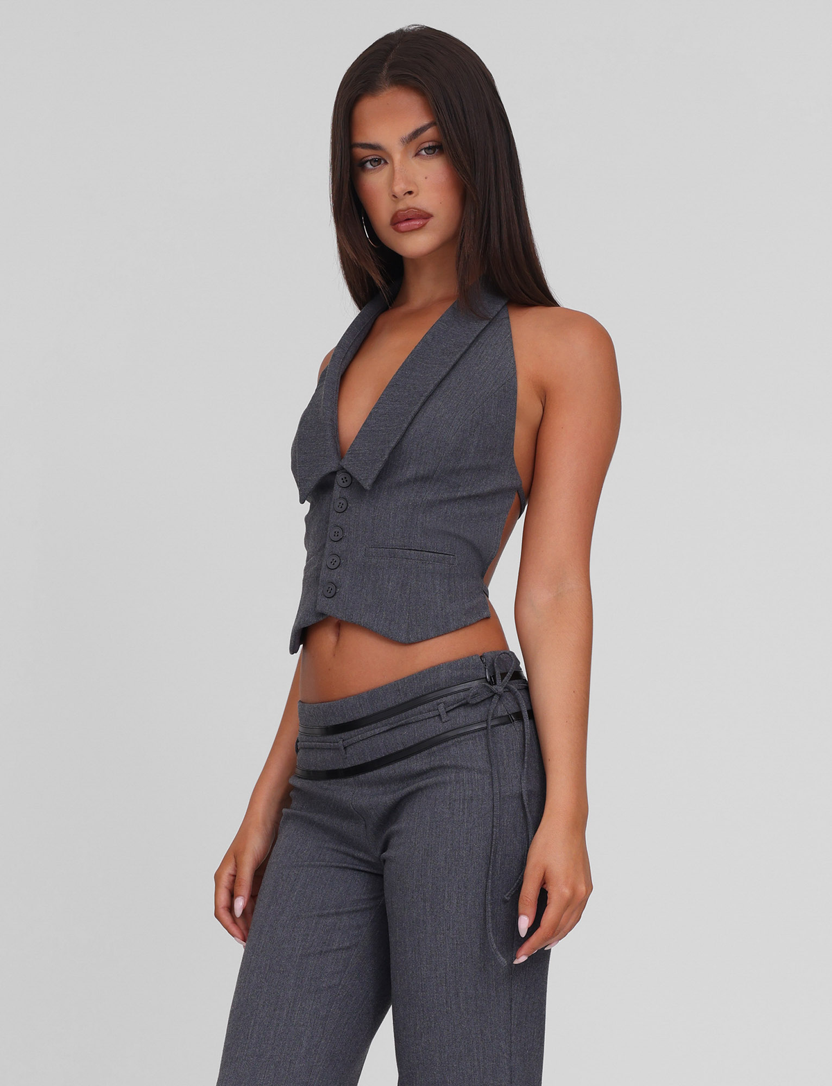 A woman with long dark hair wears the TIGERMIST FEDRA VEST - GREY, a tailored sleeveless deep V-neck vest with buttons, paired with matching gray pants featuring side-tie details, standing against a plain light background.