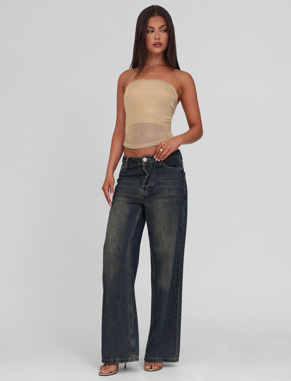 A woman with long straight brown hair and a neutral expression stands against a plain background wearing the TIGERMIST HARRIETT TUBE TOP in gold, high-waisted wide-leg dark blue jeans, and heeled sandals.