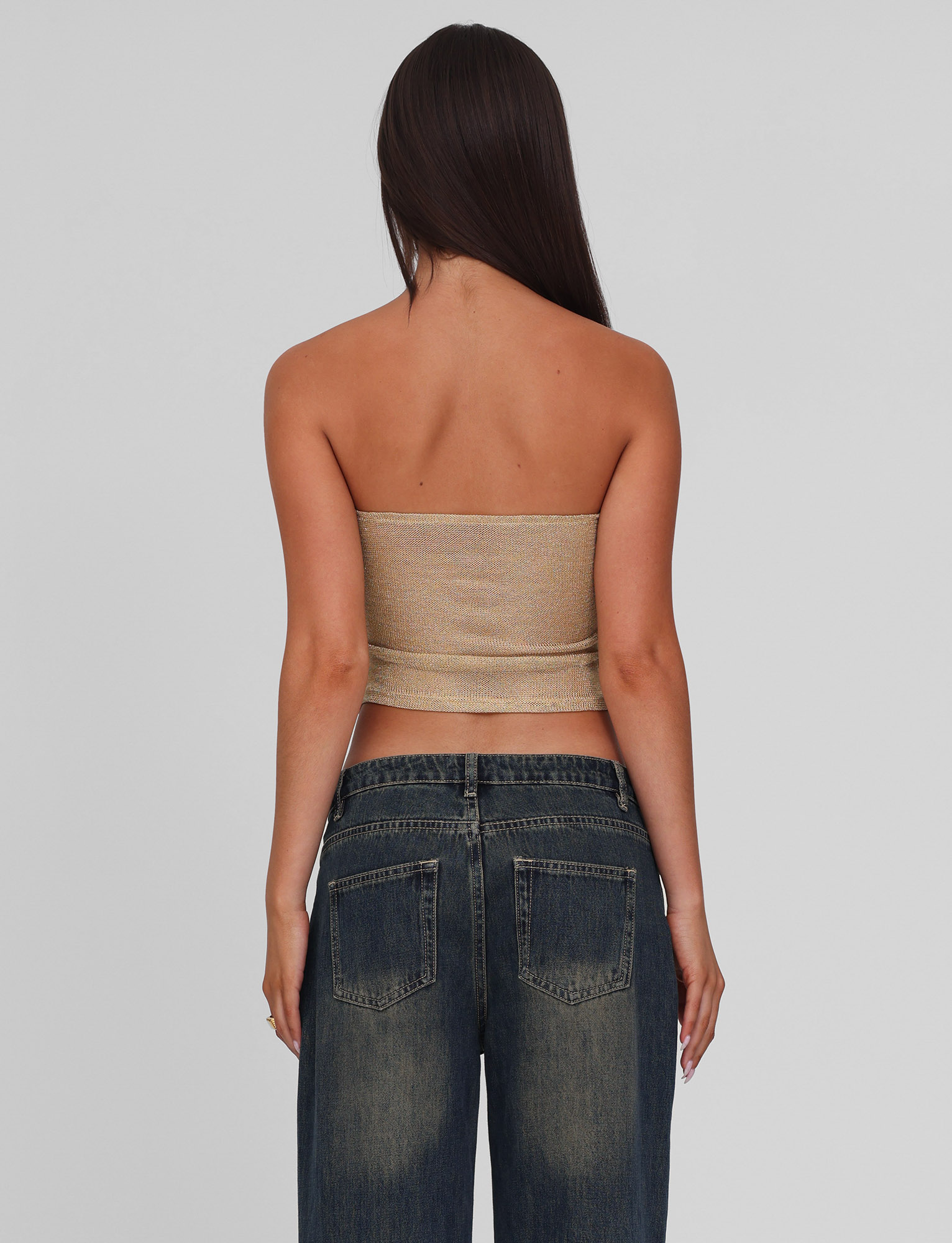 A woman with long brown hair stands facing away, wearing the TIGERMIST HARRIETT TUBE TOP in gold and dark blue jeans against a plain light gray background.