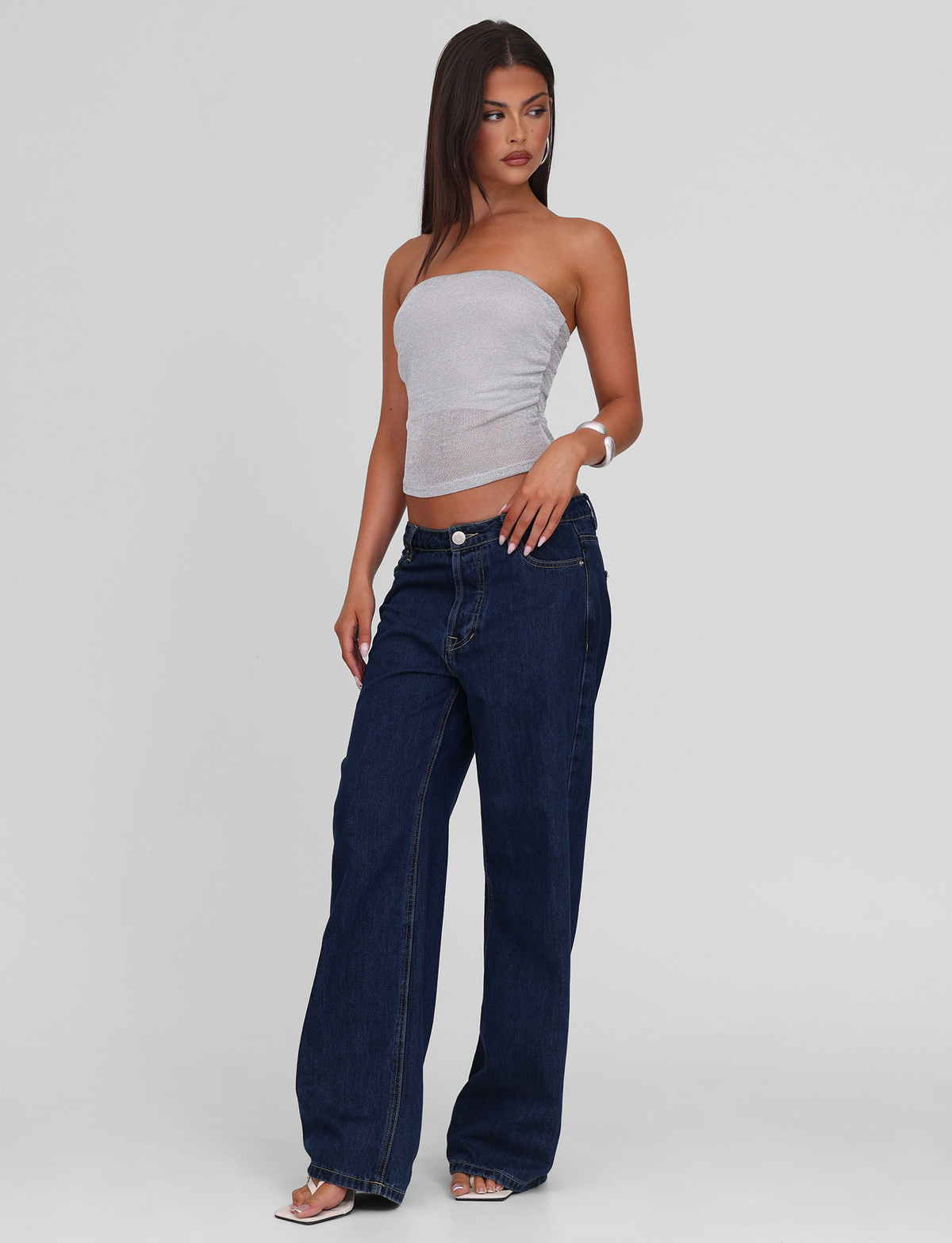 A woman with long straight hair stands against a plain background, wearing the TIGERMIST HARRIETT TUBE TOP in silver, paired with loose dark blue jeans and sandals. She looks slightly to the side with one hand by her side.