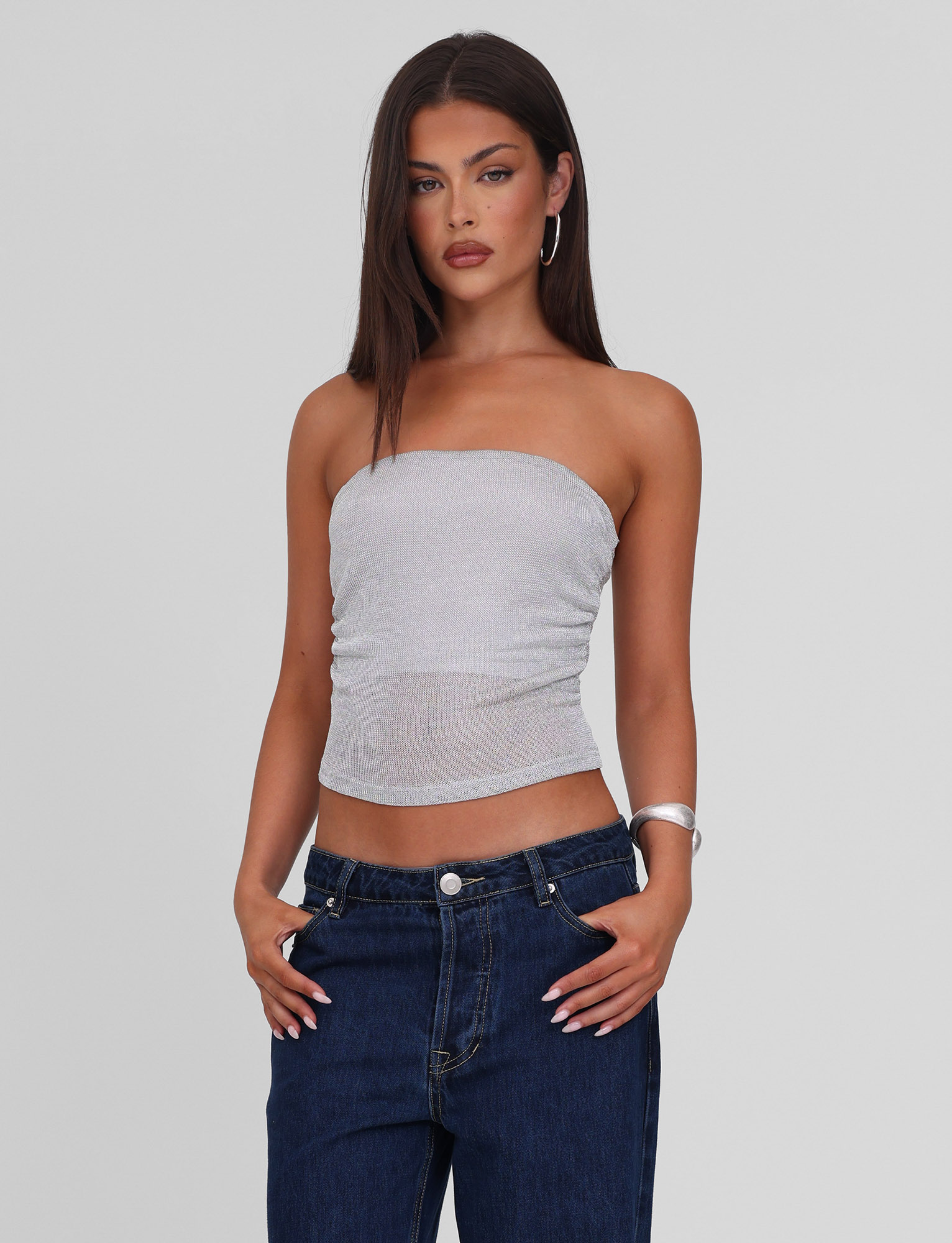 A woman with straight dark hair wears the TIGERMIST HARRIETT TUBE TOP in silver, paired with dark blue jeans, silver hoop earrings, and a silver bangle. She stands against a light gray background with her hands in her pockets.