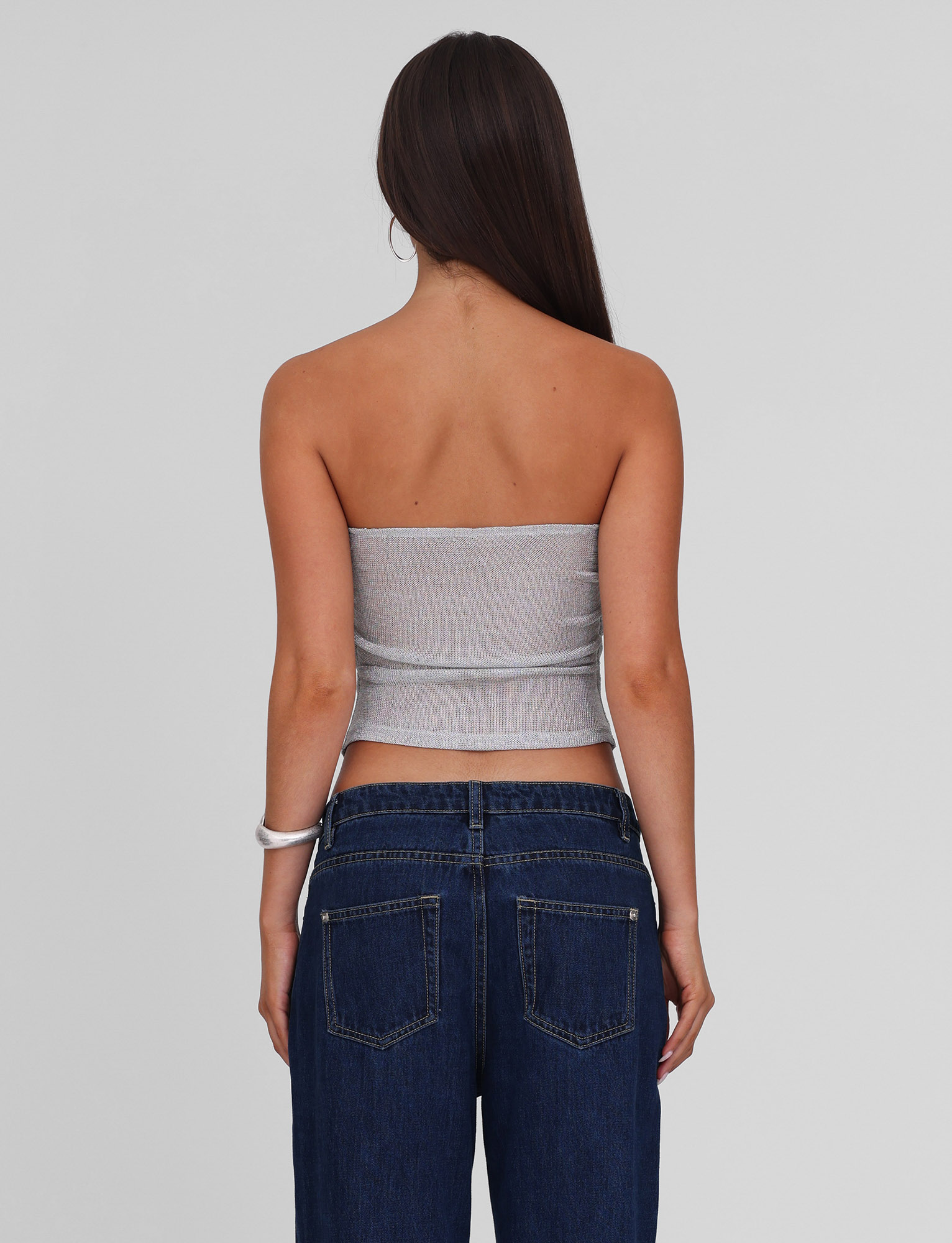 A woman with long straight brown hair is seen from behind, wearing the TIGERMIST HARRIETT TUBE TOP in silver and dark blue jeans against a plain light grey background.
