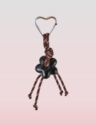 The Tigermist MIRIAM KEYCHAIN - CHOCOLATE features a heart-shaped metal carabiner clasp, attached to dark brown rope knotted decoratively. Four knotted ends dangle a black flower charm, set against a light pink background for soft contrast.