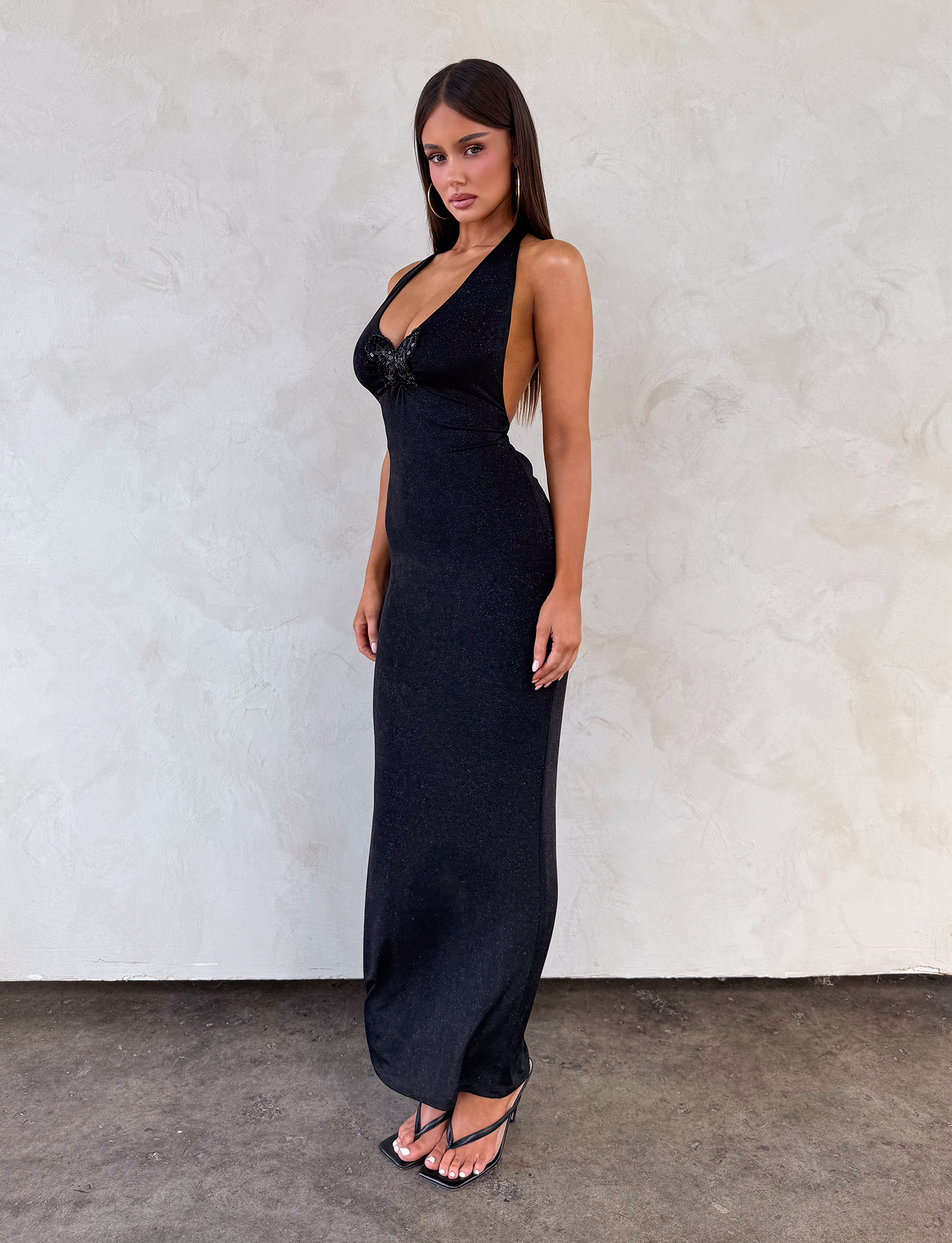 A woman with long dark hair models the TIGERMIST ASTORIA MAXI DRESS - BLACK, a sleeveless glitter maxi dress, paired with black sandals as she poses against a light textured wall.