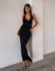 A woman with long dark hair stands against a white wall, wearing the TIGERMIST ASTORIA MAXI DRESS in black with a halter neckline and black strappy sandals, creating a minimalist, modern vibe.