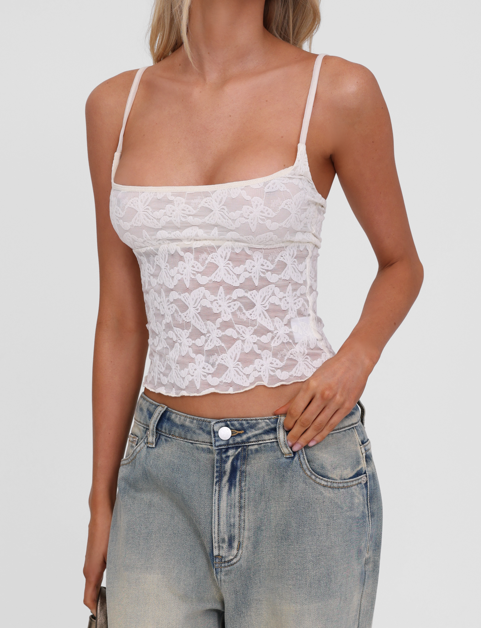 A woman wears the TIGERMIST GIULIA 2PC TOP in ivory lace with adjustable straps and a butterfly pattern, paired with light blue high-waisted jeans, standing against a plain background with one hand in her pocket.