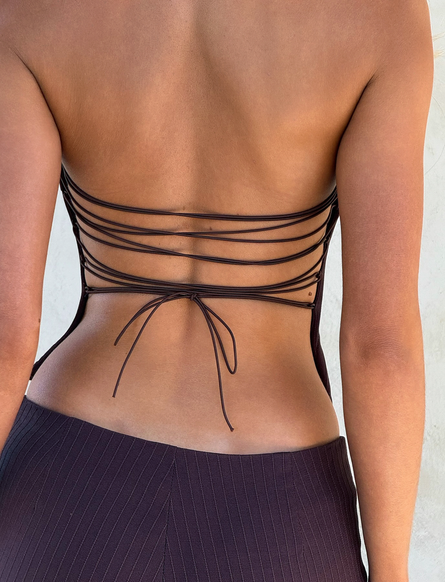 A person wears the TIGERMIST GWENDOLINE TOP in chocolate, a backless brown top with thin crisscross straps tied in a bow, paired with brown pinstriped pants.