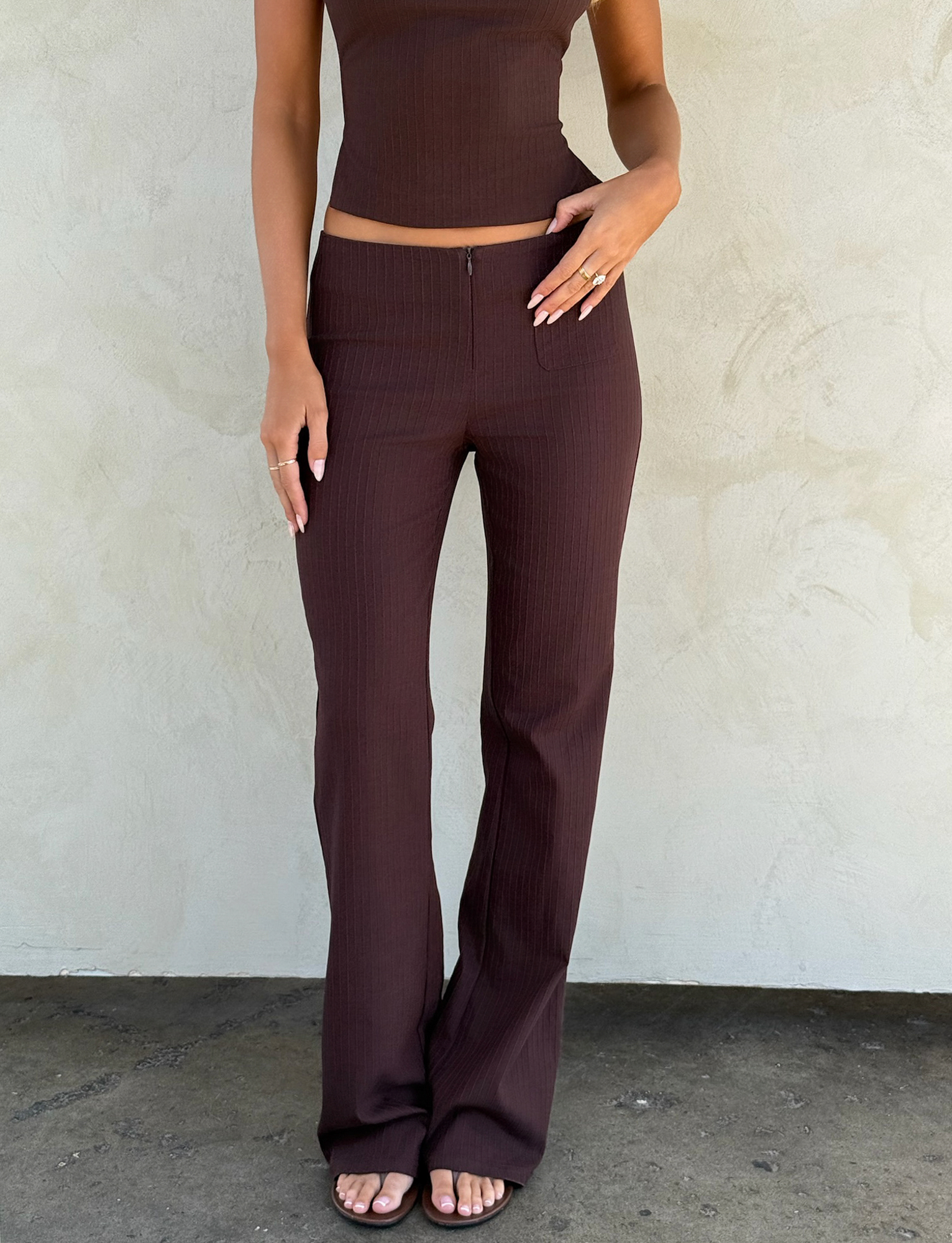 A woman stands against a textured light wall, wearing TIGERMIST's GWENDOLINE PANT in chocolate with a flared leg and a sleeveless fitted top, one hand on her waist and the other by her side.