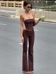 A woman wearing TIGERMIST's GWENDOLINE PANT in chocolate, with flared legs and a matching strapless top, stands on a city street. She holds a green drink, sports sunglasses and an updo, and carries a small brown shoulder bag.