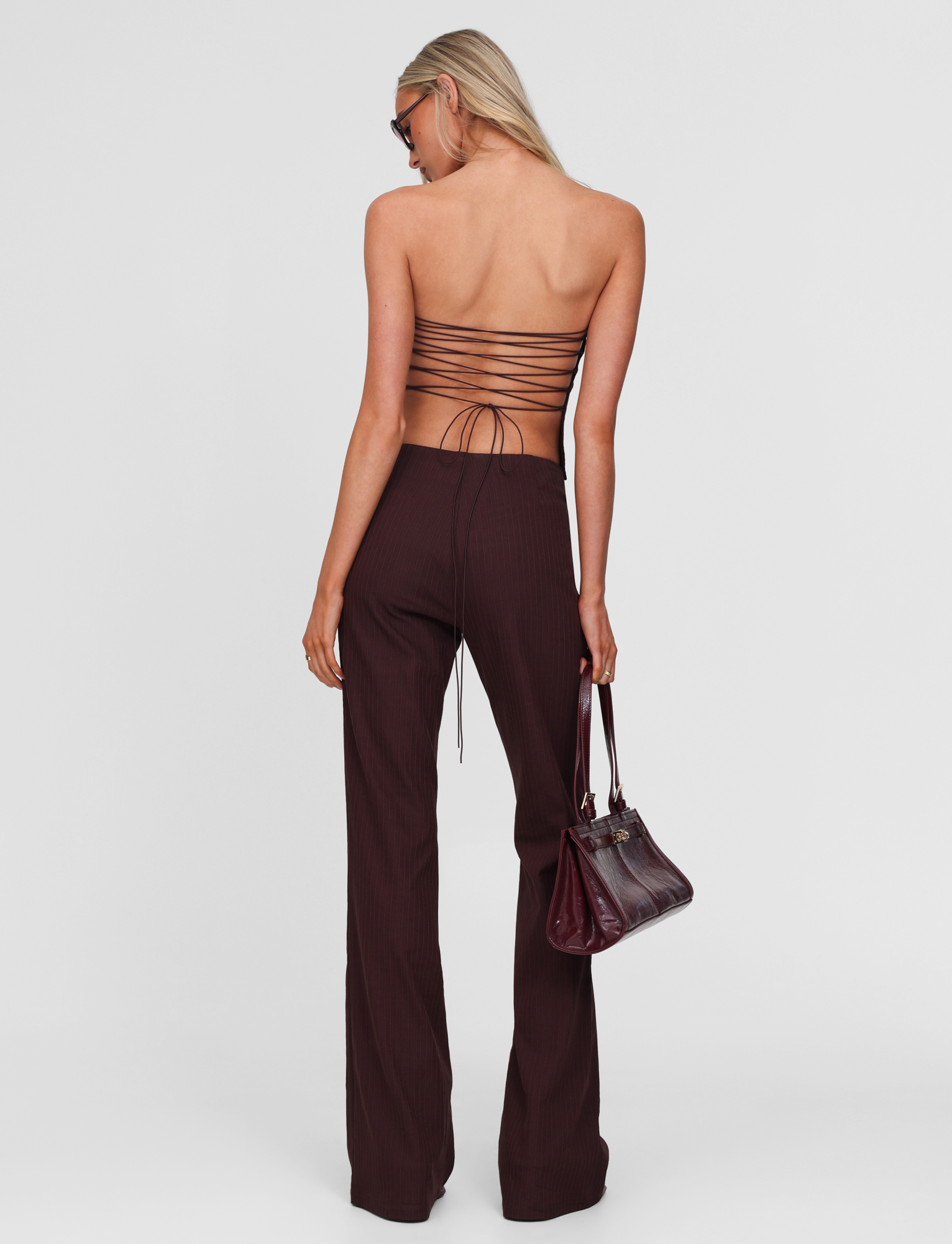 A woman with long blonde hair, wearing TIGERMIST’s GWENDOLINE PANT in chocolate with flared legs and a strappy backless top, holds a dark burgundy handbag and wears sunglasses against a plain white background.