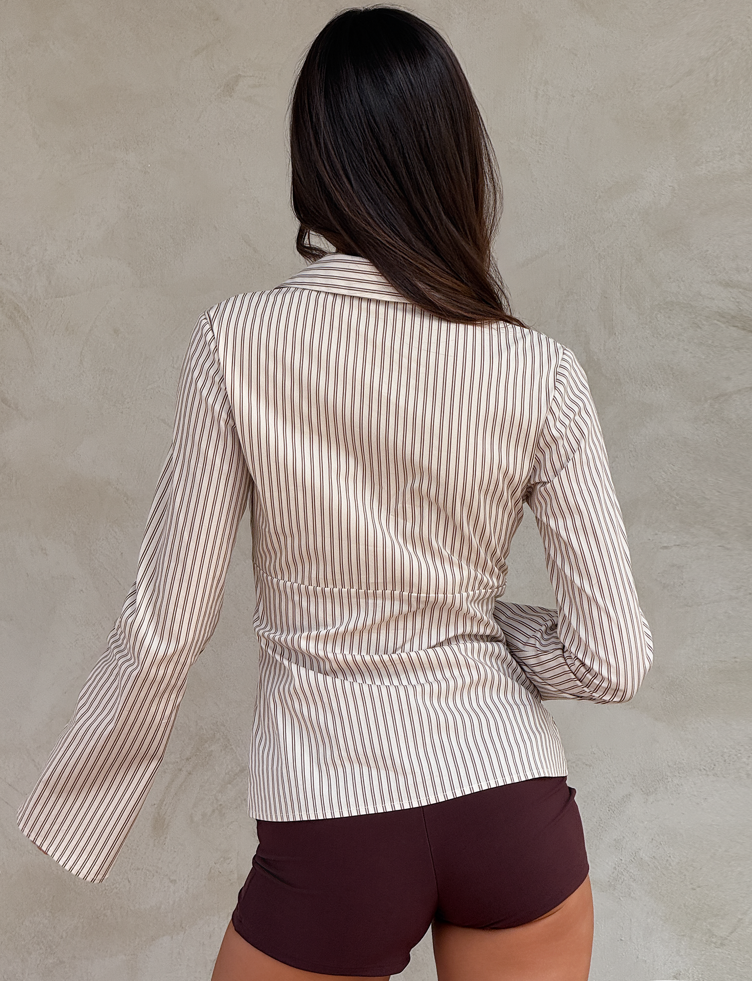 A woman with long brown hair stands facing a textured beige wall, her back to the camera. She wears the TIGERMIST SABRINE SHIRT - BEIGE STRIPE styled with dark brown shorts.