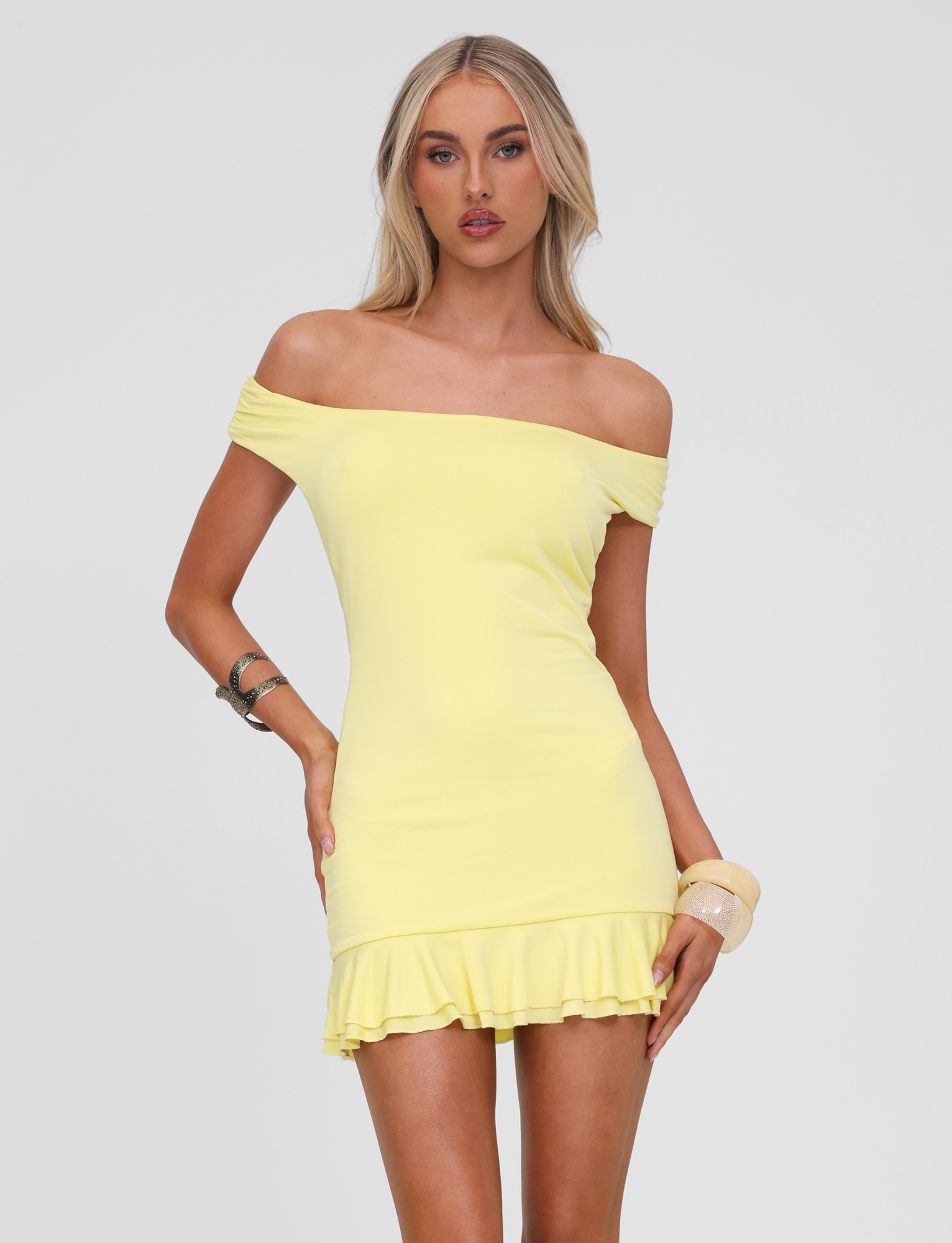 A woman poses against a white background wearing the TIGERMIST CLIA MINI DRESS in lemon, an off-shoulder mini dress with a frill hemline. She has long blonde hair and accessorizes with bracelets on both wrists.
