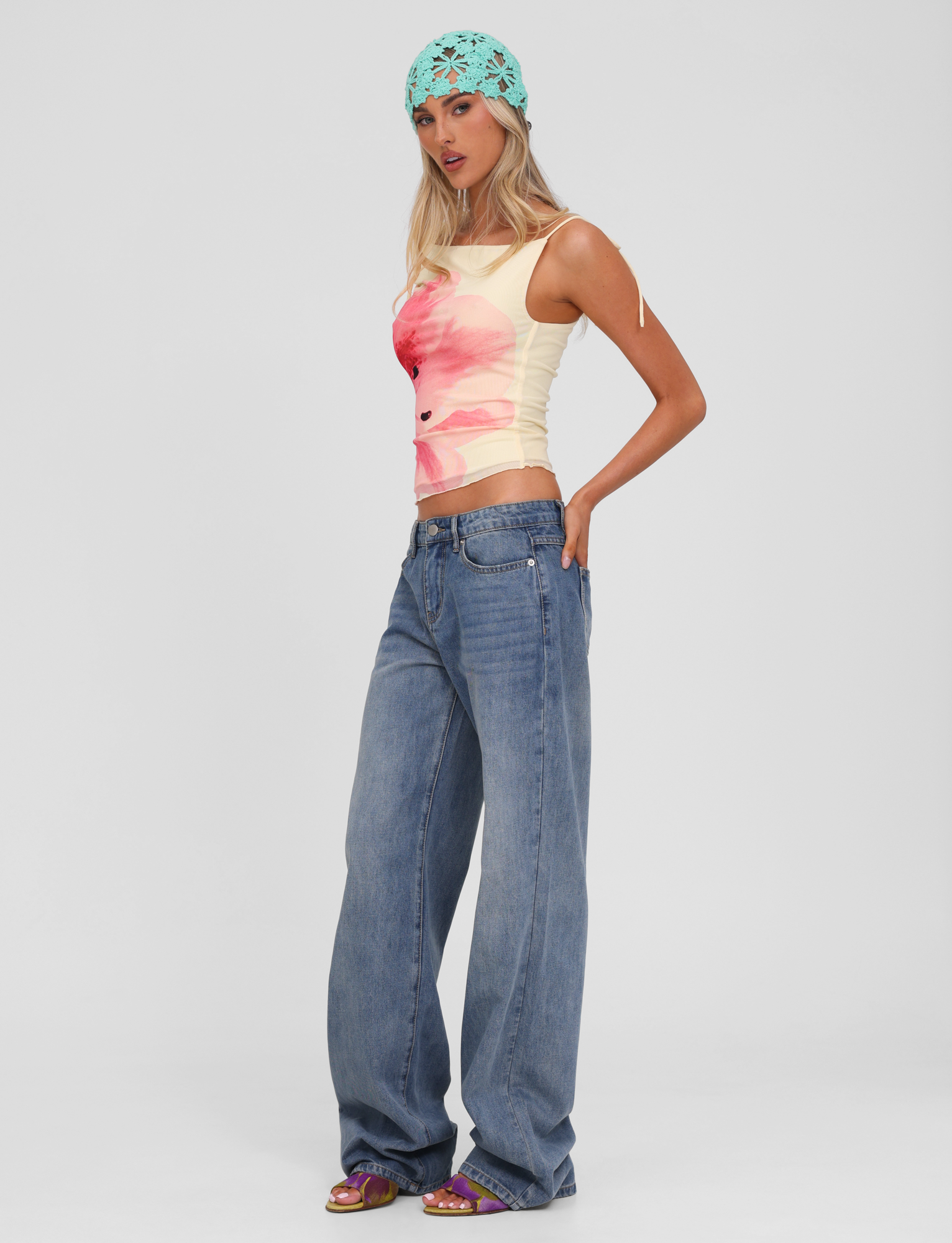 A woman poses against a white background wearing TIGERMIST's ESTRELLA JEAN - MID WASH, a fitted sleeveless top with a bold pink flower print, purple sandals, and an aqua blue crocheted headscarf.