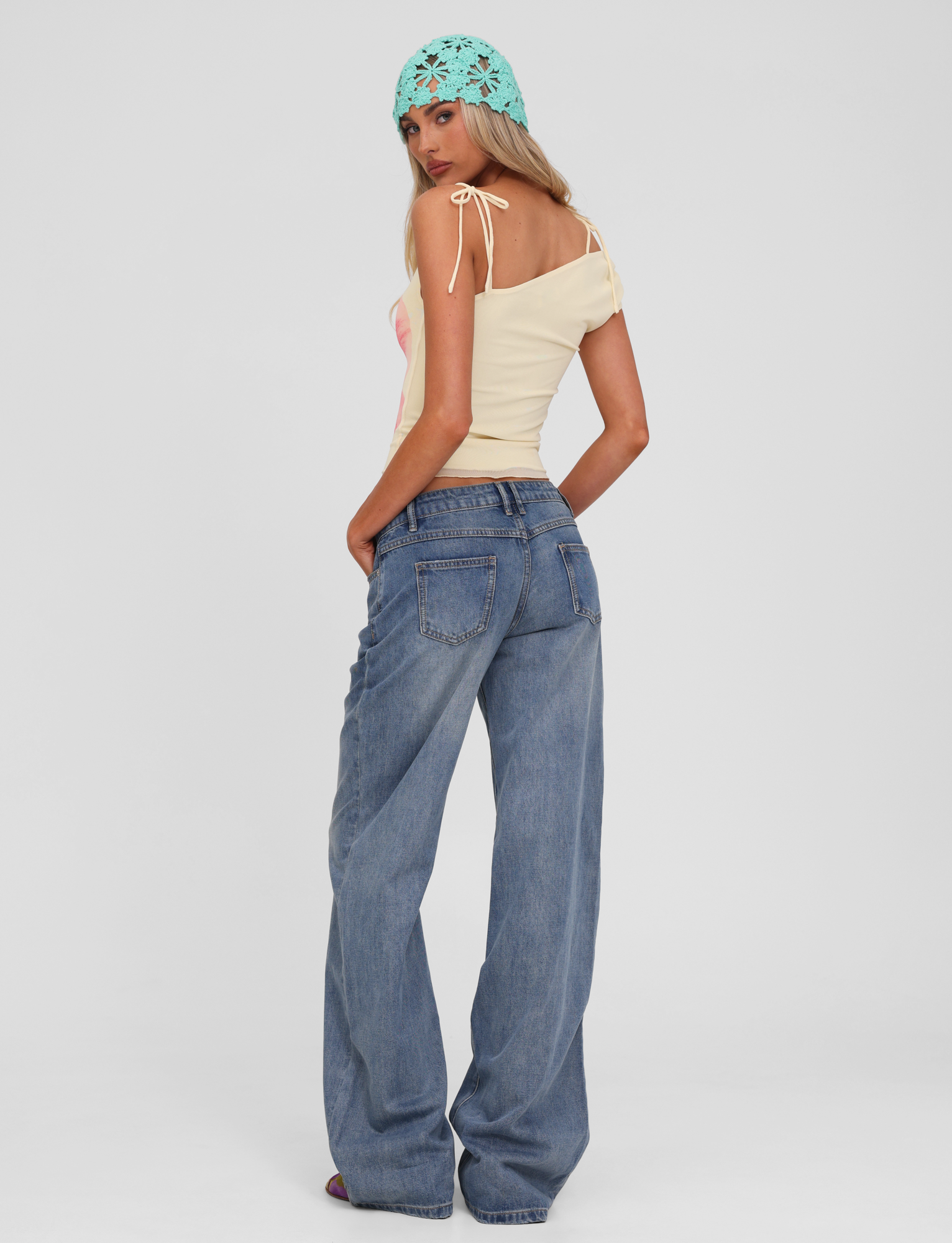 A woman stands against a white background, looking over her shoulder. She wears TIGERMIST’s ESTRELLA JEAN - MID WASH, a yellow off-shoulder top, and a light blue crochet headscarf.