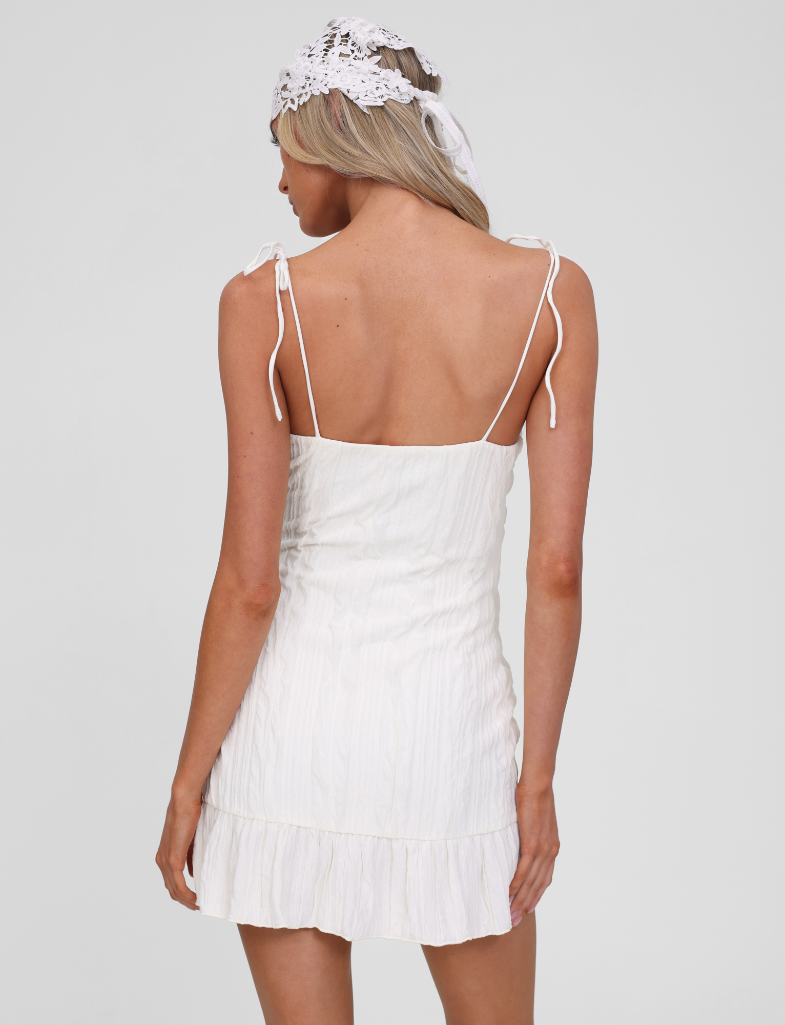 A woman with long blonde hair wears the TIGERMIST RANIA MINI DRESS in off white, featuring tie shoulder straps and a frill hemline. She pairs it with a white lace headpiece, standing with her back to the camera against a plain background.