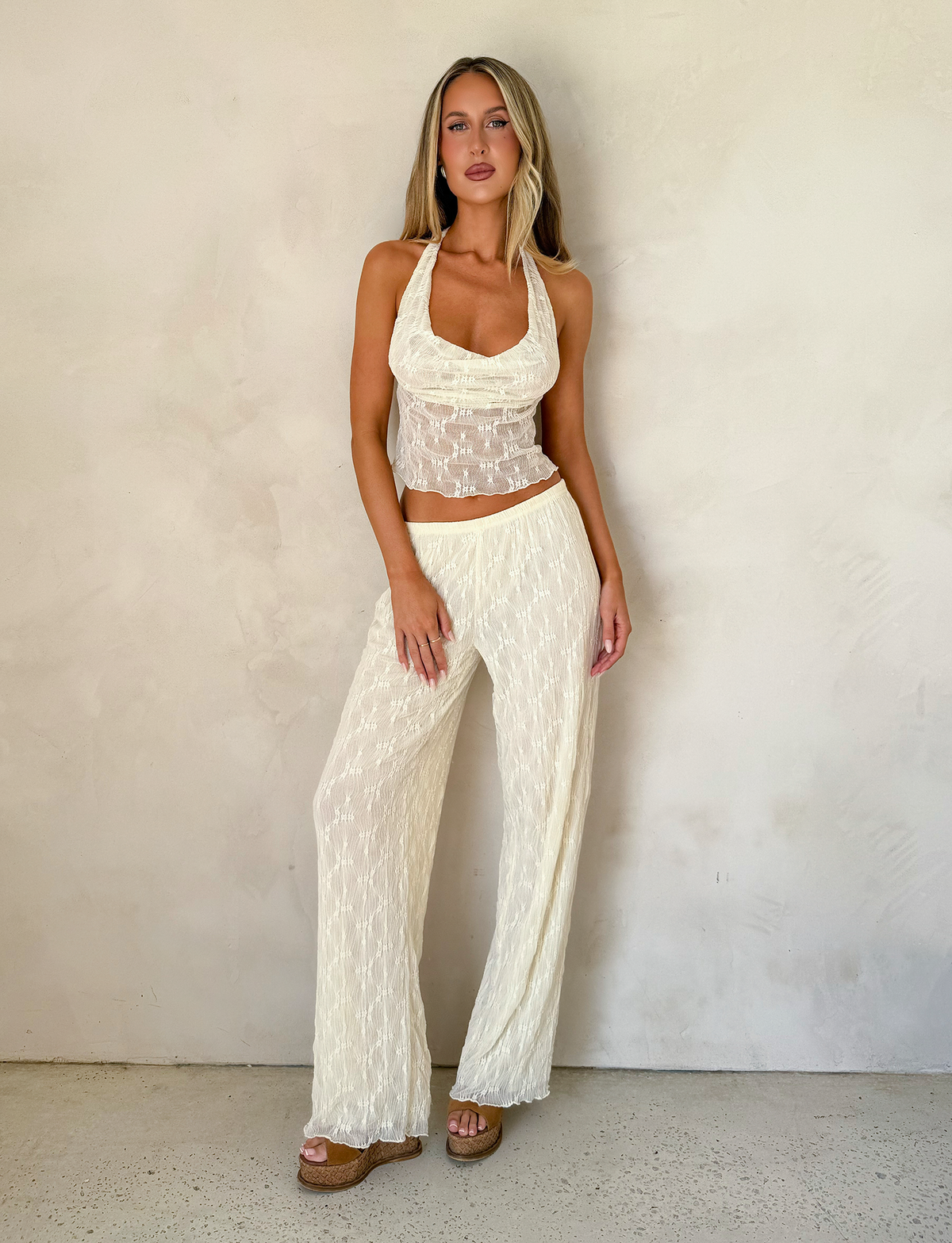 A woman with long, straight hair stands by a light wall, wearing the TIGERMIST KATERINA TOP in ivory lace with matching wide-leg pants and platform sandals.