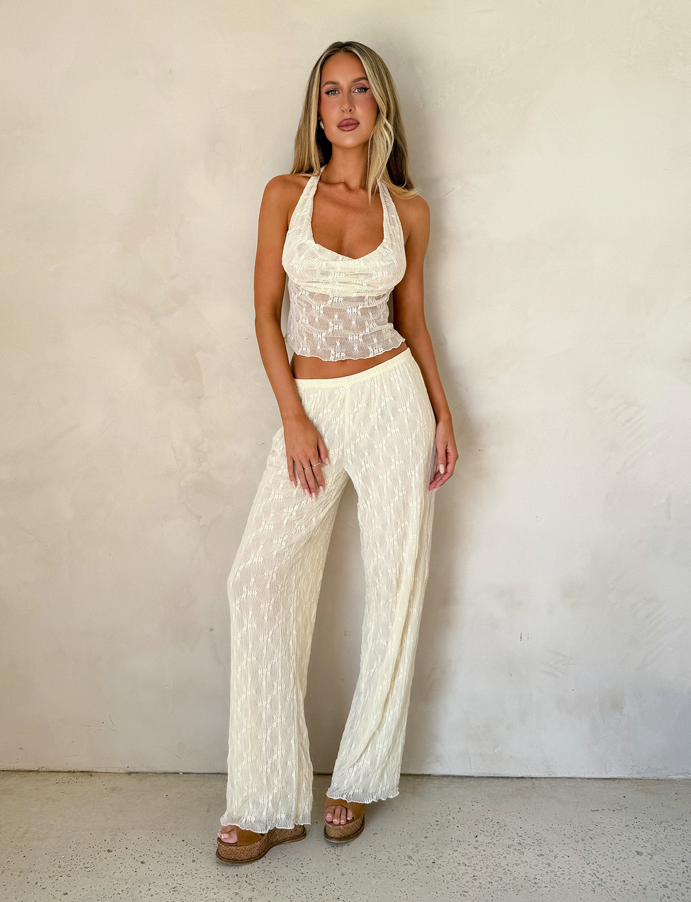 A woman stands against a light wall, wearing TIGERMIST's KATERINA PANT in ivory lace with a subtle textured pattern, paired with an ivory semi-sheer halter top and platform sandals. Her long hair is worn loose.