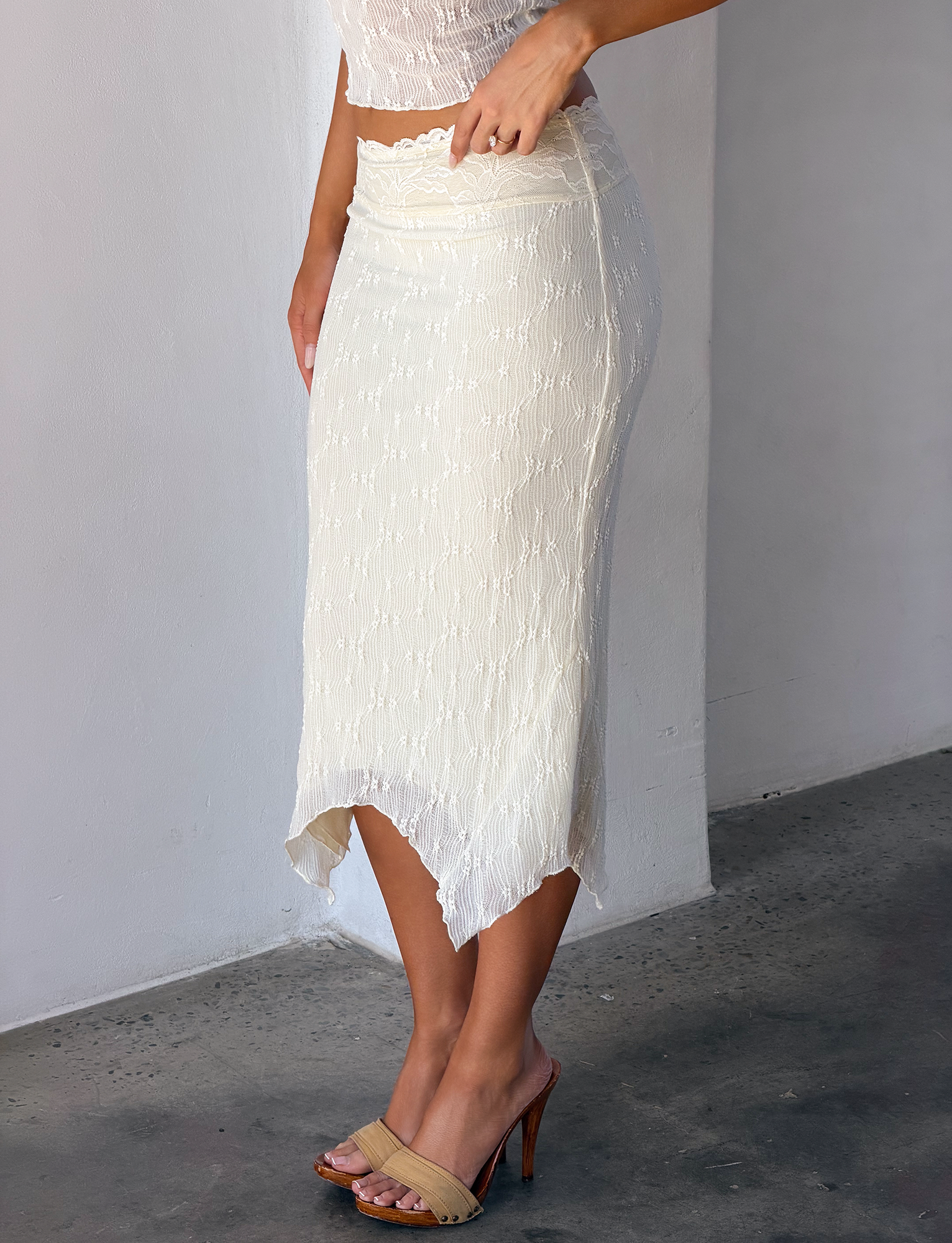 A person wears the TIGERMIST KATERINA MIDI SKIRT in cream, featuring stretch lace and an asymmetrical hem, with a matching crop top and tan high-heeled sandals, standing indoors on a concrete floor.