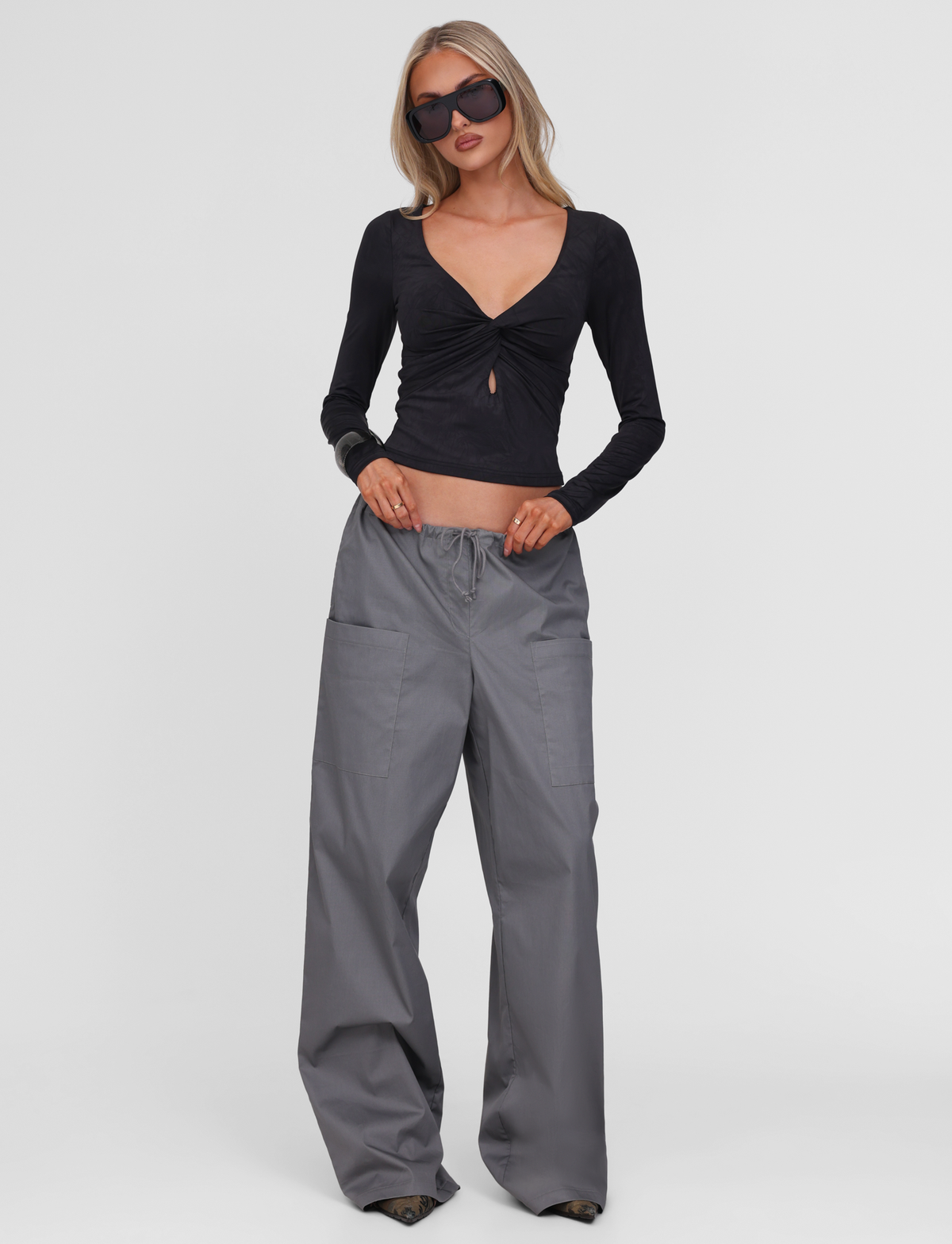 A woman wears sunglasses, gray cargo pants, and the TIGERMIST SAFFIRA LONG SLEEVE TOP in black, featuring a front twist and keyhole detail, standing against a plain white background.