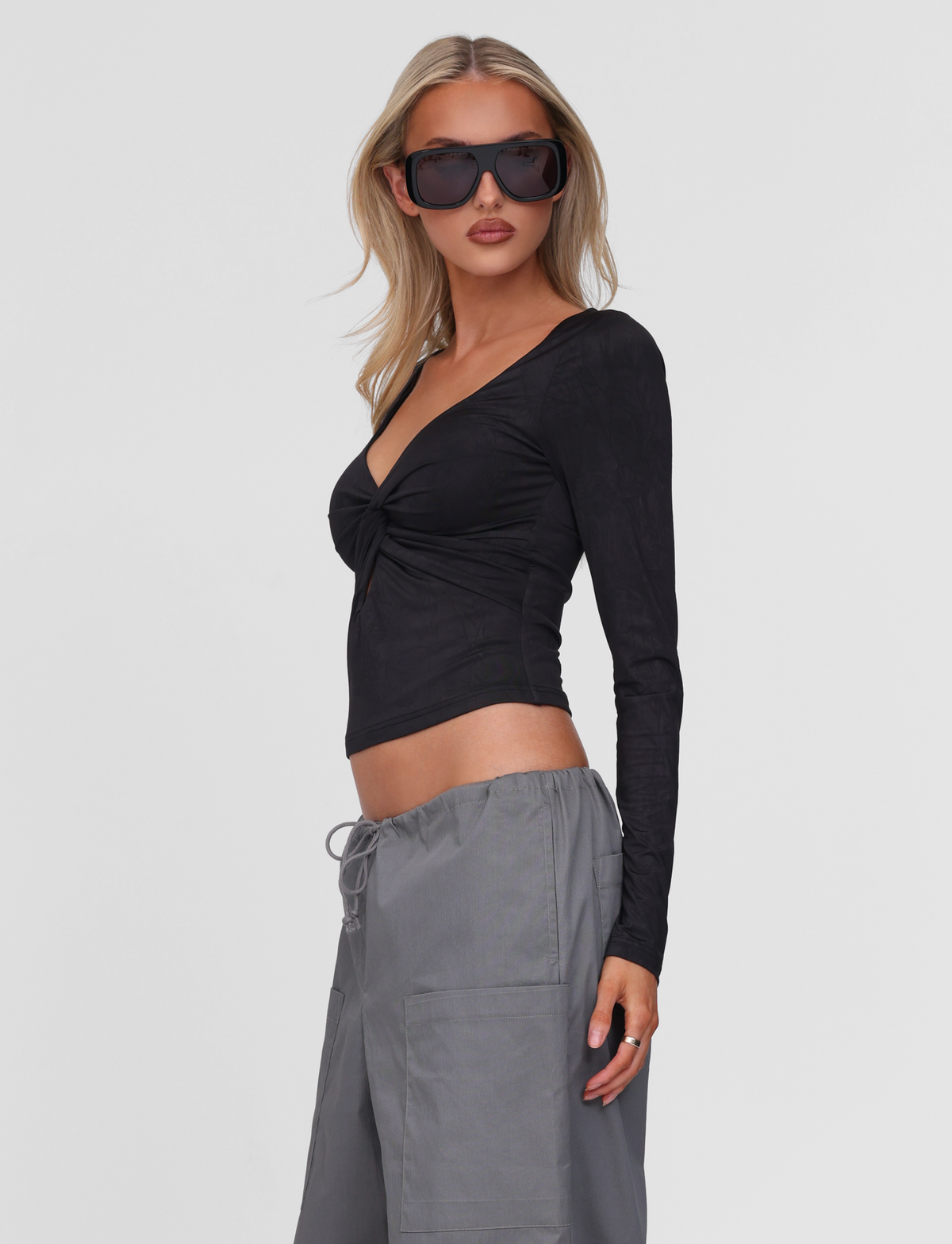 A woman with long blonde hair wears oversized black sunglasses, loose gray pants, and the TIGERMIST SAFFIRA LONG SLEEVE TOP in black, standing against a plain white background and looking confidently to the side.