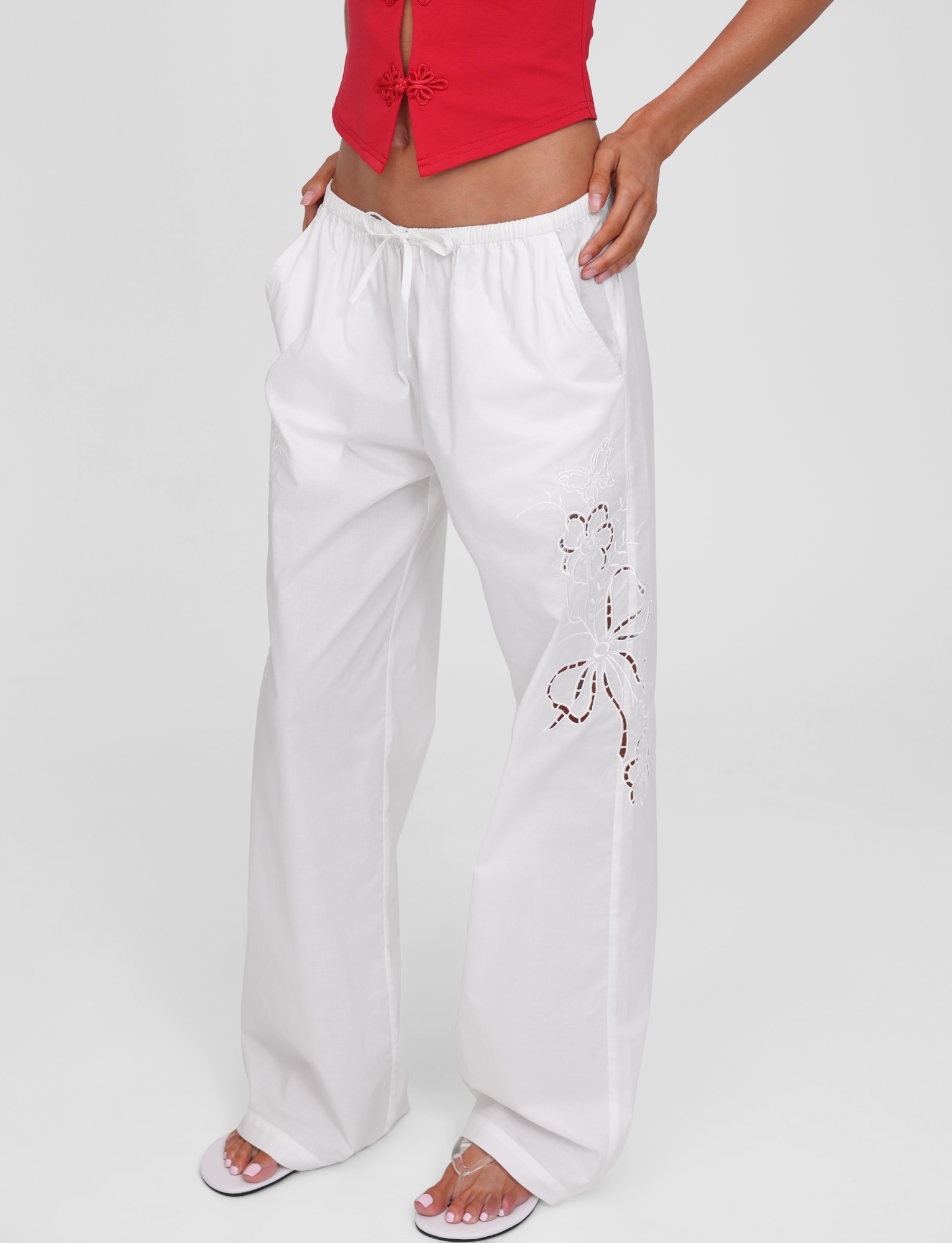 A person wears the TIGERMIST CRESSY PANT in white—loose, wide-leg cotton with floral embroidery on one leg—paired with a red sleeveless top and white sandals, standing hands-in-pockets against a plain background.