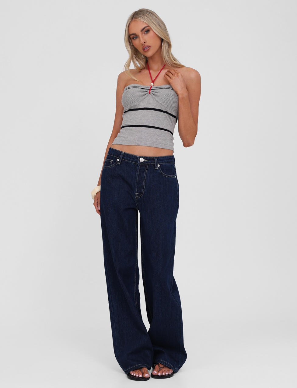 A woman poses against a plain background in the TIGERMIST NOUR TOP - GREY STRIPE and loose dark blue jeans, with long blonde hair and one hand touching her neck.