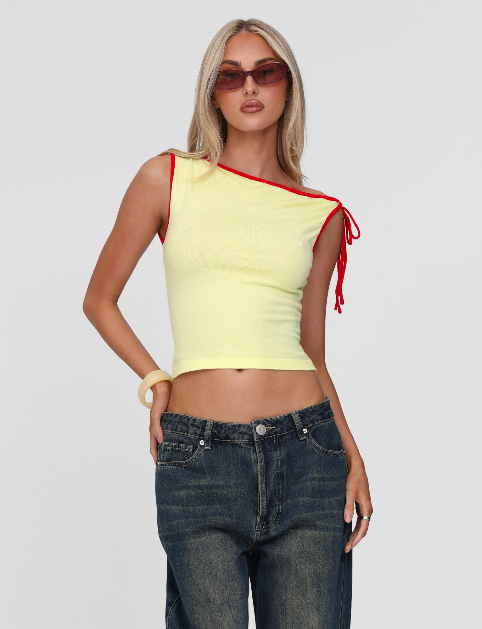 A woman in the TIGERMIST ALIRA TOP - YELLOW, featuring red trim and a tie detail on one shoulder, sunglasses, dark loose jeans, and a chunky bracelet, poses against a plain light background.