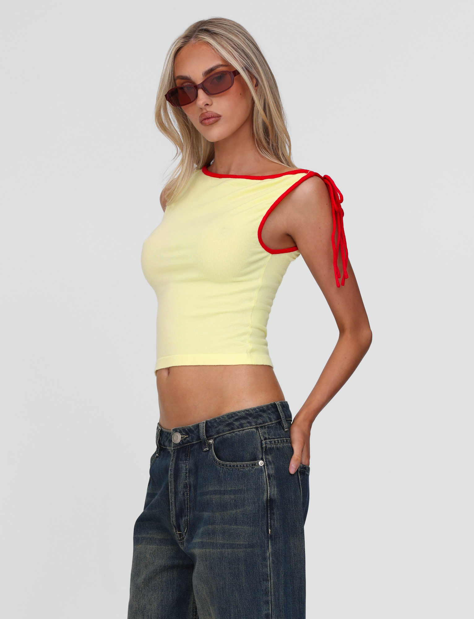 A woman wears the TIGERMIST ALIRA TOP in yellow—sleeveless, off-the-shoulder with red trim and a tie detail—pairing it with blue jeans and sunglasses, standing against a plain light background.