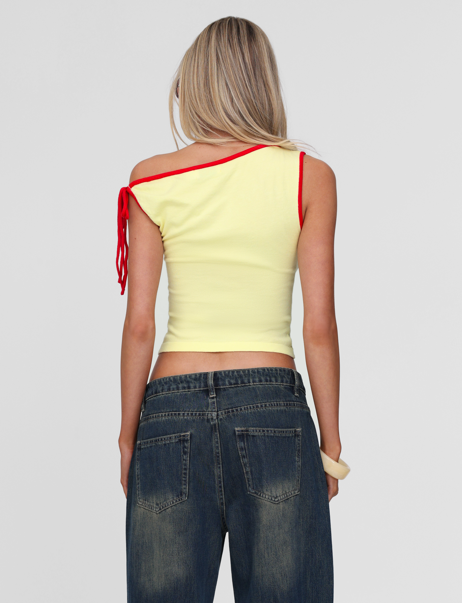 A woman with long blonde hair faces away from the camera against a light background, wearing the TIGERMIST ALIRA TOP in yellow with red trim, styled off the shoulder with an asymmetrical neckline and paired with blue jeans.