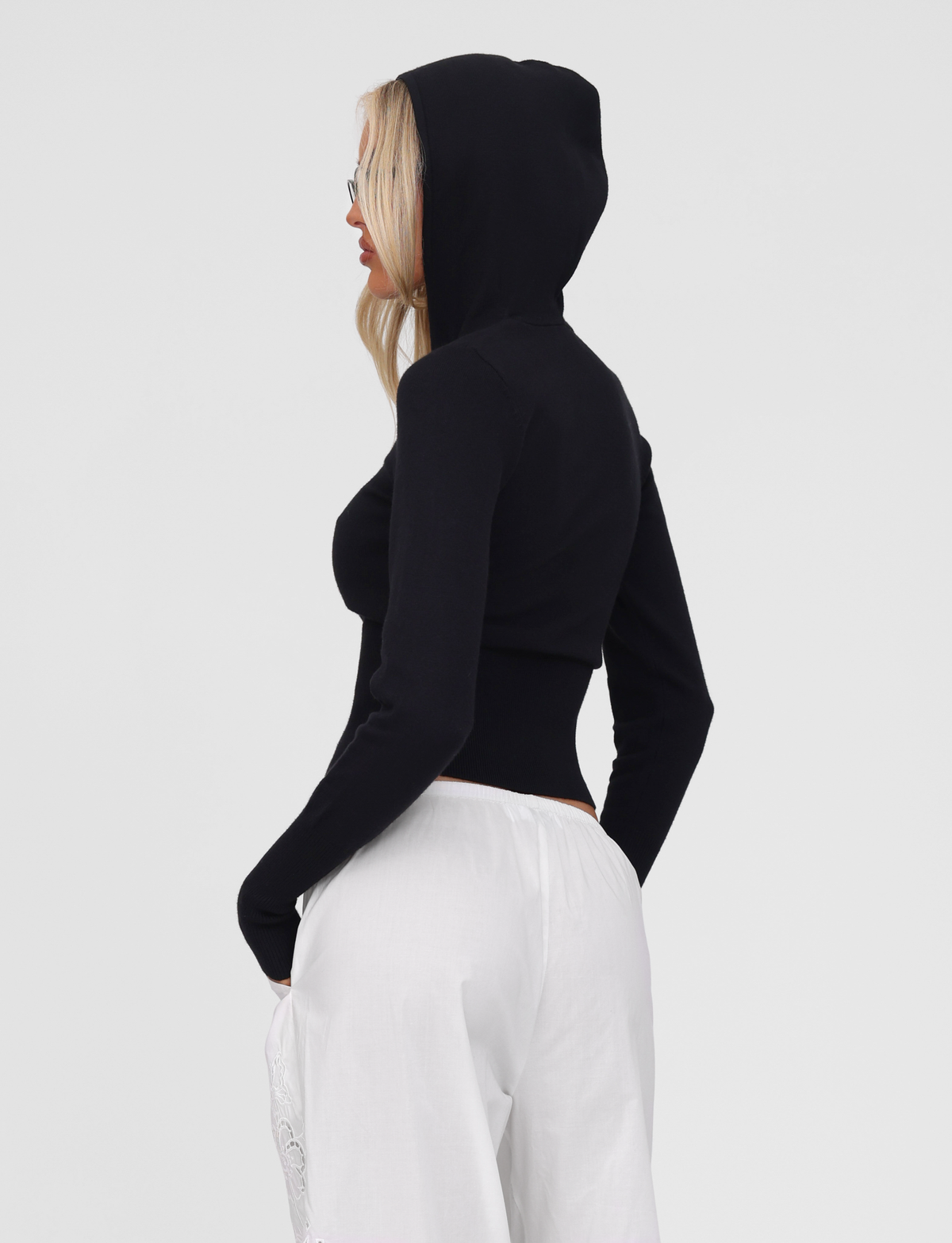A woman in profile wears the TIGERMIST SARINA JUMPER in black, featuring a gold zipper, paired with white pants. She has long blonde hair, sunglasses, and her hands are in her pockets against a plain background.