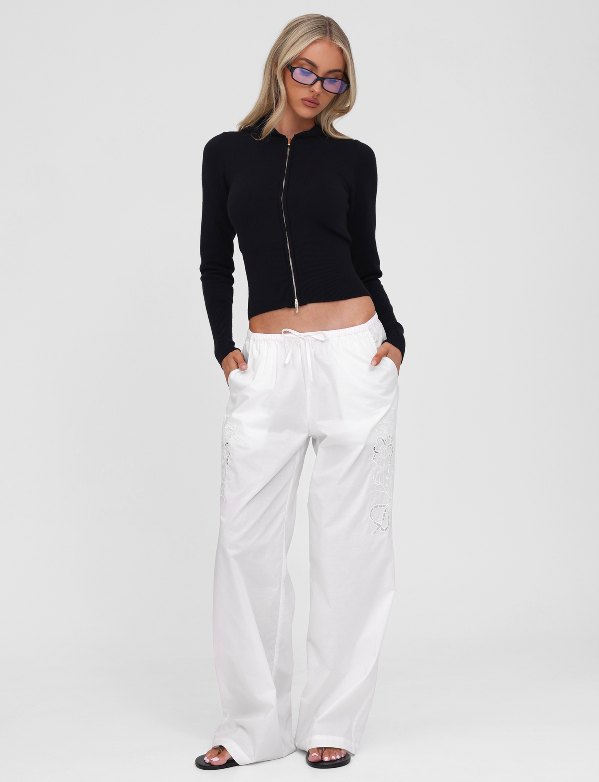 Wearing the TIGERMIST SARINA JUMPER in black, paired with loose white pants, black slides, and blue-tinted glasses, a person stands against a plain white background.