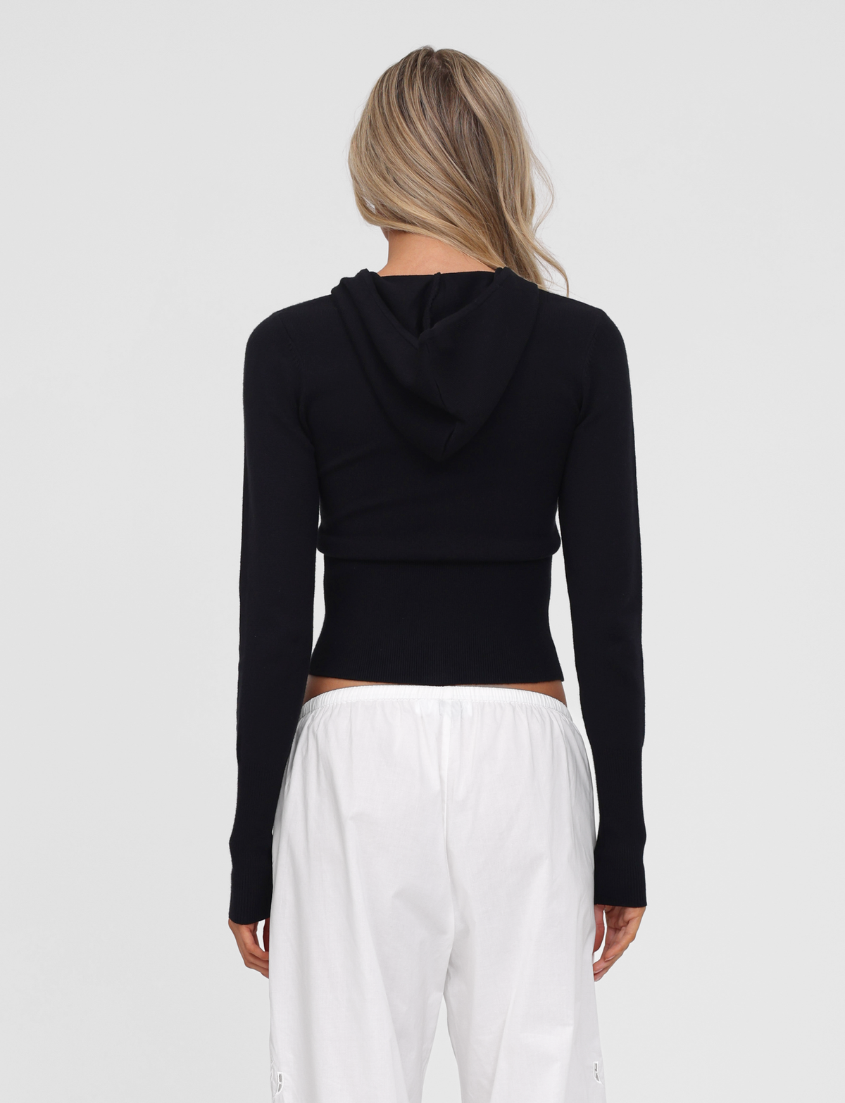 A person with long blonde hair faces away, wearing the TIGERMIST SARINA JUMPER in black and loose white pants against a plain light background.