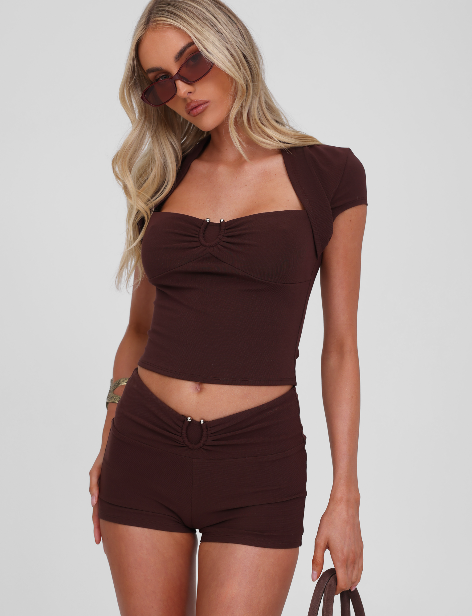 A woman with long blonde hair wears the TIGERMIST LUNARA SHORT in chocolate with a fitted dark brown crop top, sunglasses, bracelet, and bag, posing against a plain light background.