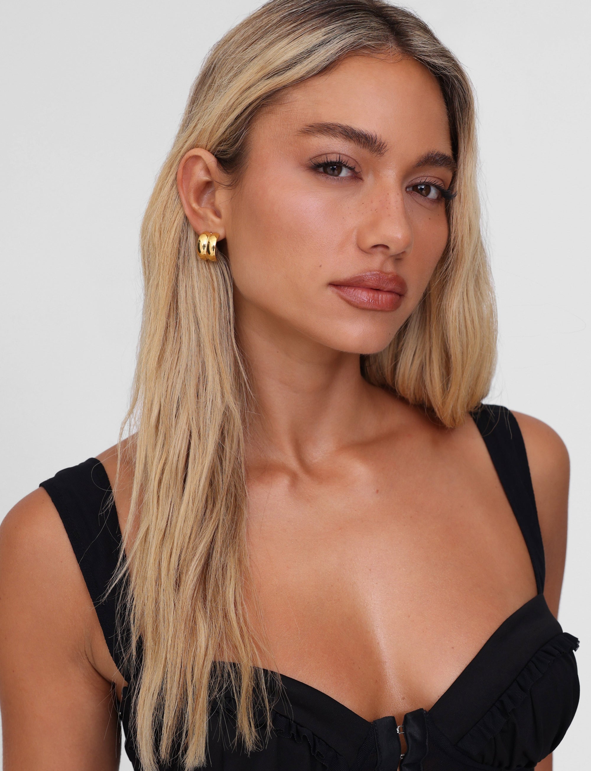 A woman with long, blonde hair poses confidently in a black top and LILLIA - GOLD earrings by TIGERMIST. She stands against a plain background, her calm expression complemented by softly framing hair.