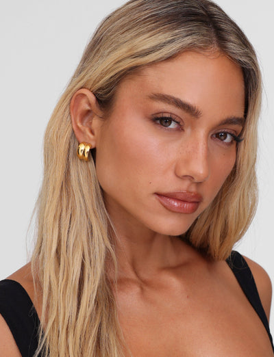 A woman with long blonde hair wears LILLIA EARRING - GOLD by TIGERMIST, gazing confidently at the camera. Her light makeup and neutral lip color stand out against a plain background, and her black thin-strapped top complements her striking accessories perfectly.