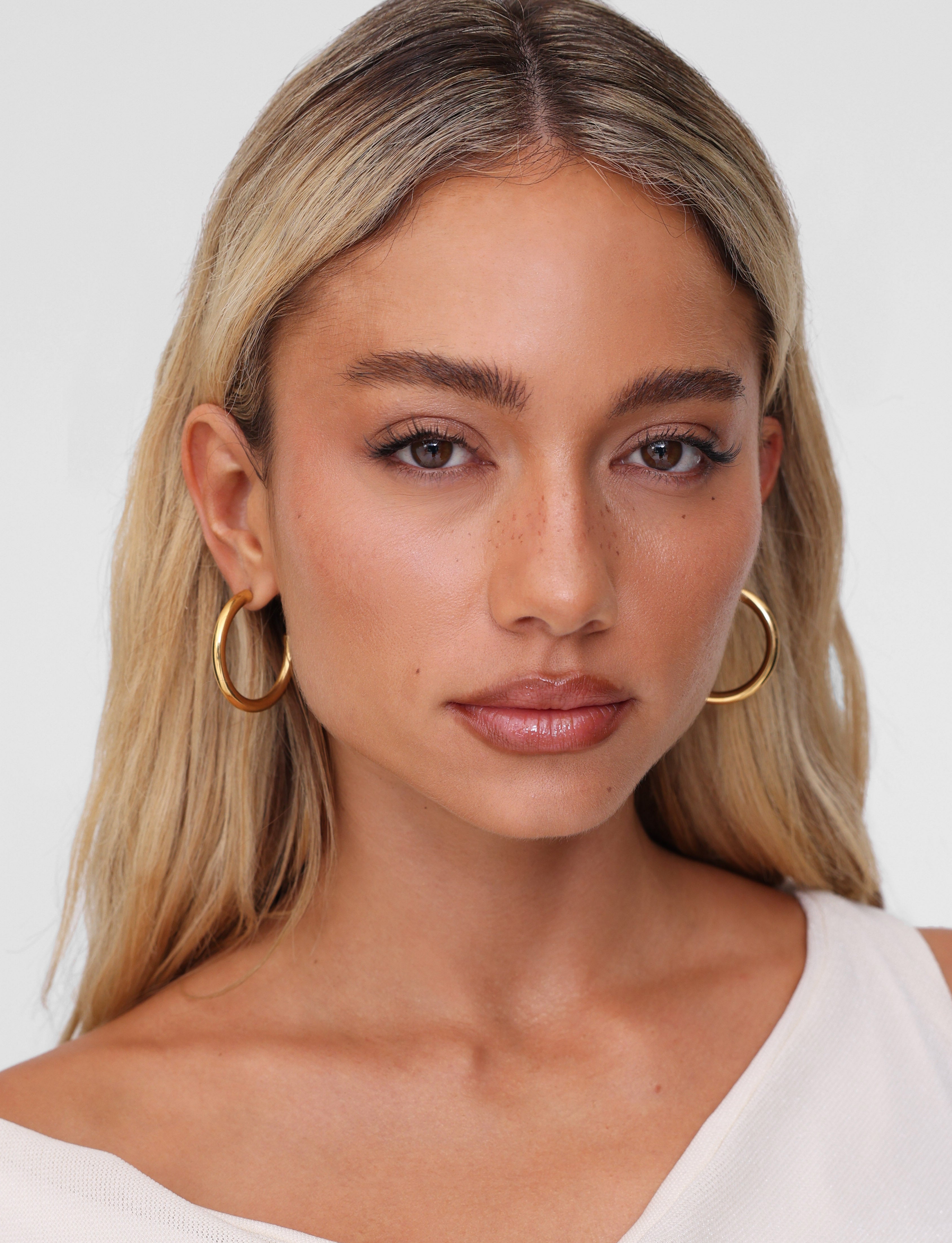 A person with long blond hair and the lightweight PIPER EARRING - GOLD by TIGERMIST gazes directly at the camera, wearing an off-the-shoulder, light-colored top against a plain, light background.
