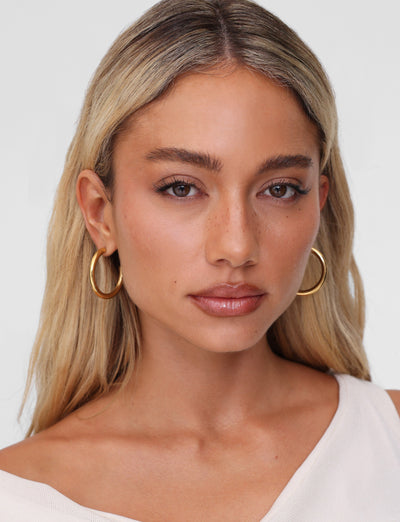 A person with long blond hair and the lightweight PIPER EARRING - GOLD by TIGERMIST gazes directly at the camera, wearing an off-the-shoulder, light-colored top against a plain, light background.