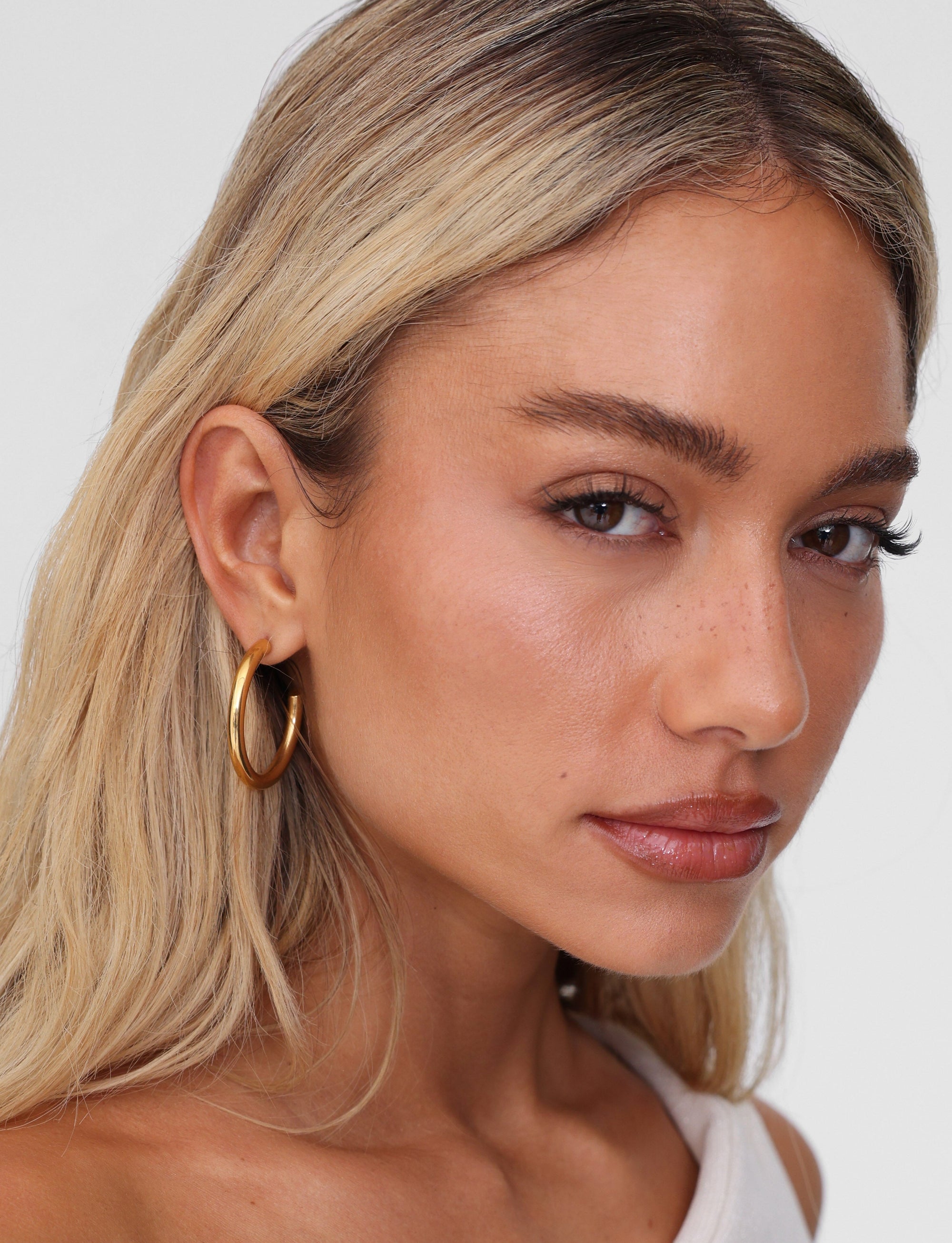 A woman with long blonde hair and PIPER EARRING - GOLD by TIGERMIST gazes at the camera. Her white top complements subtle natural makeup, and her earrings are hypoallergenic and lightweight.