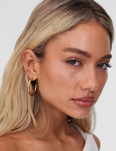 A woman with long blonde hair and PIPER EARRING - GOLD by TIGERMIST gazes at the camera. Her white top complements subtle natural makeup, and her earrings are hypoallergenic and lightweight.