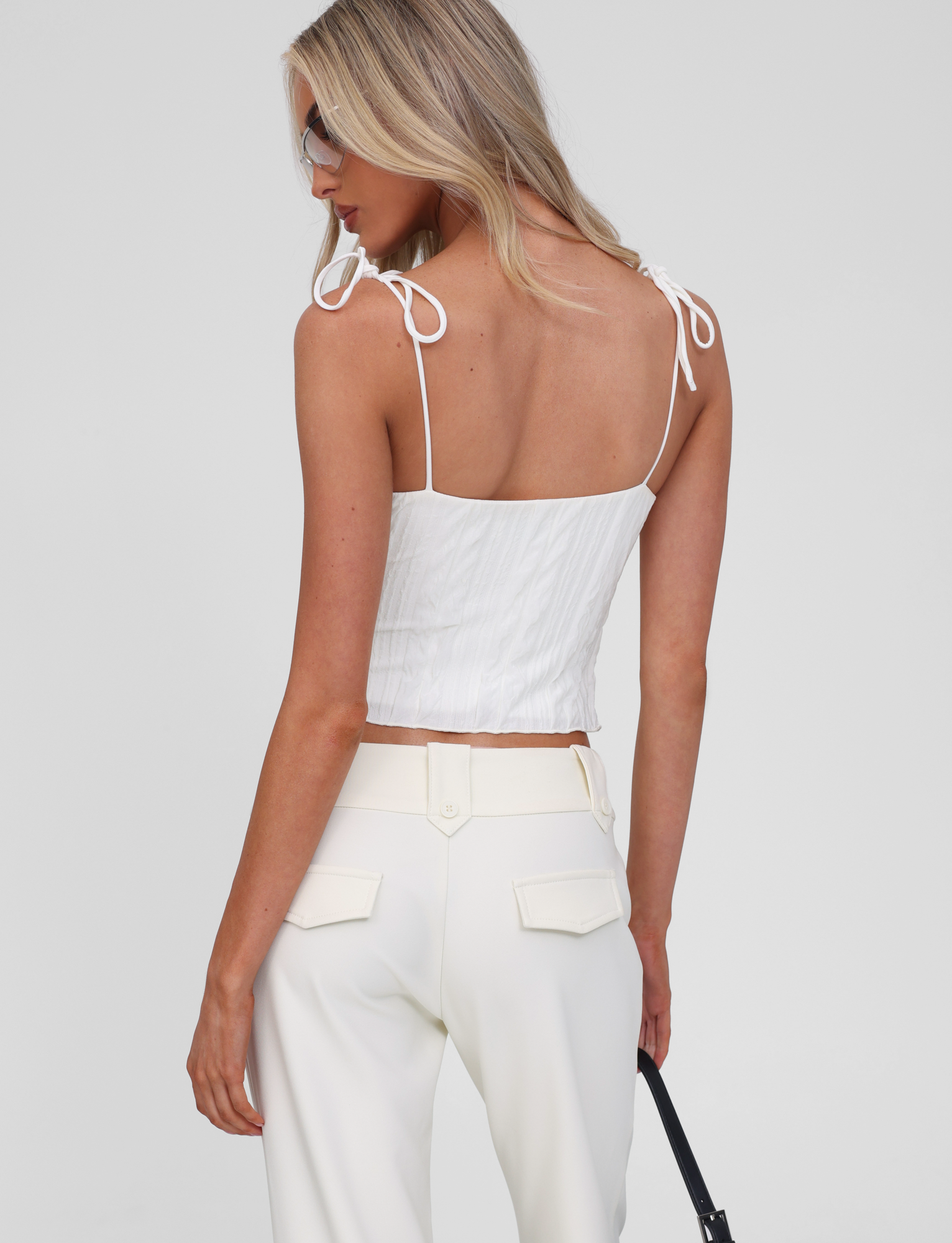 A woman with long blonde hair models the TIGERMIST RANIA TOP in off white, featuring strappy shoulder details and a sleeveless design. She faces away, looks over her shoulder, and holds a black strap against a plain background.