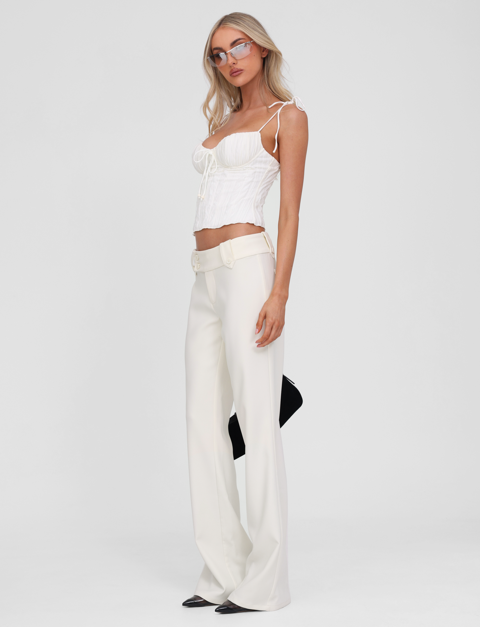 A woman stands against a plain white background wearing the TIGERMIST RANIA TOP in off white, styled with high-waisted wide-leg white pants, black pointy shoes, glasses, and holding a black clutch behind her.