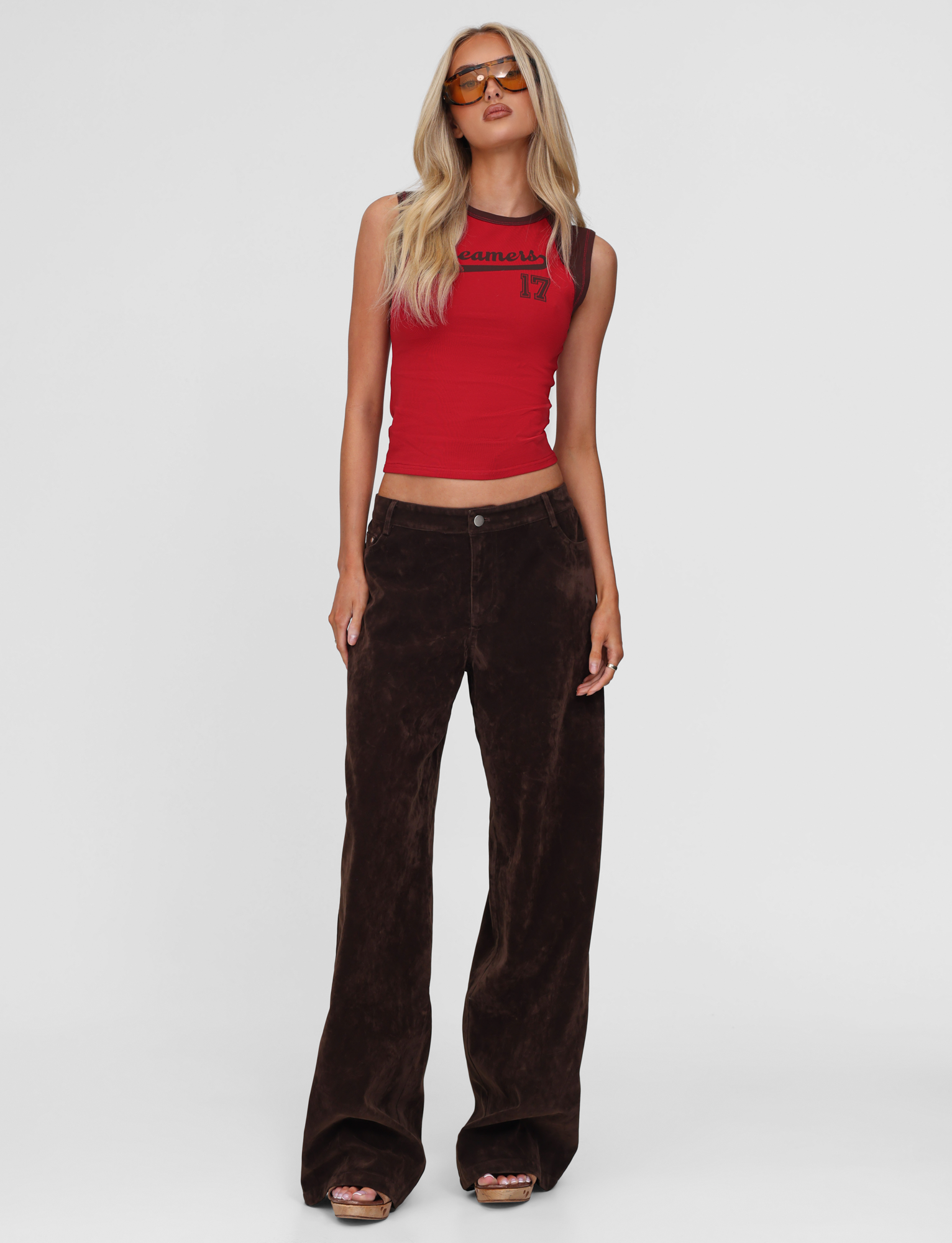 A woman with long blonde hair models the TIGERMIST FIERA TEE in red, paired with loose dark brown velvet pants, brown platform sandals, and orange-tinted sunglasses against a plain white background.